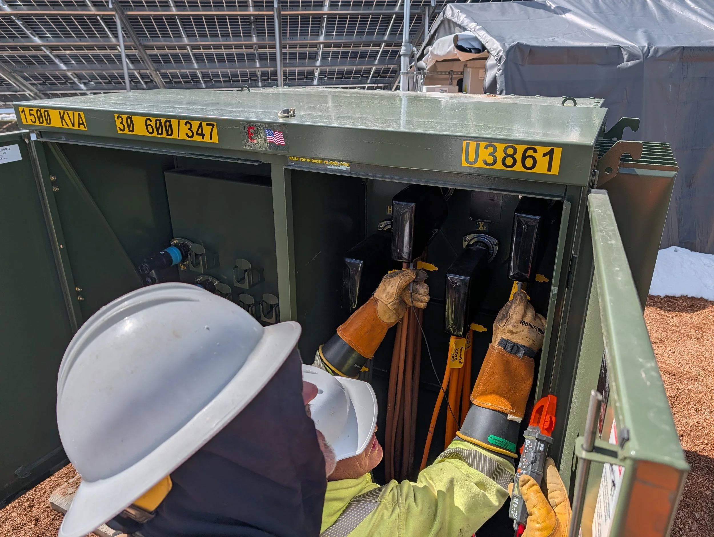 Electric utility worker wearing safety helmet and gloves installing or repairing electrical equipment inside a green metal cabinet outdoors, with solar panels on a structure overhead.