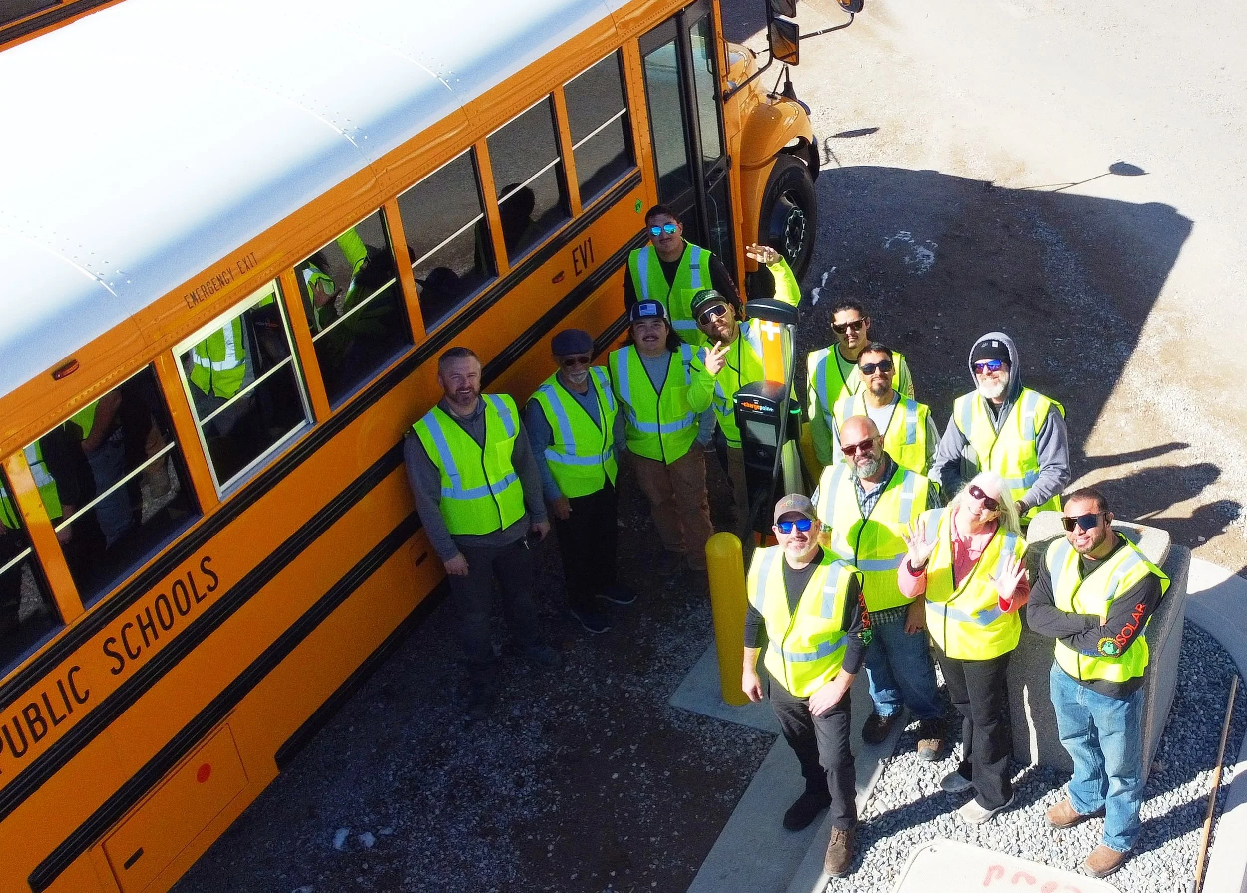 A group of people wearing yellow safety vests gathered outdoors near a yellow school bus and electric vehicle charging station, some waving and smiling at the camera.