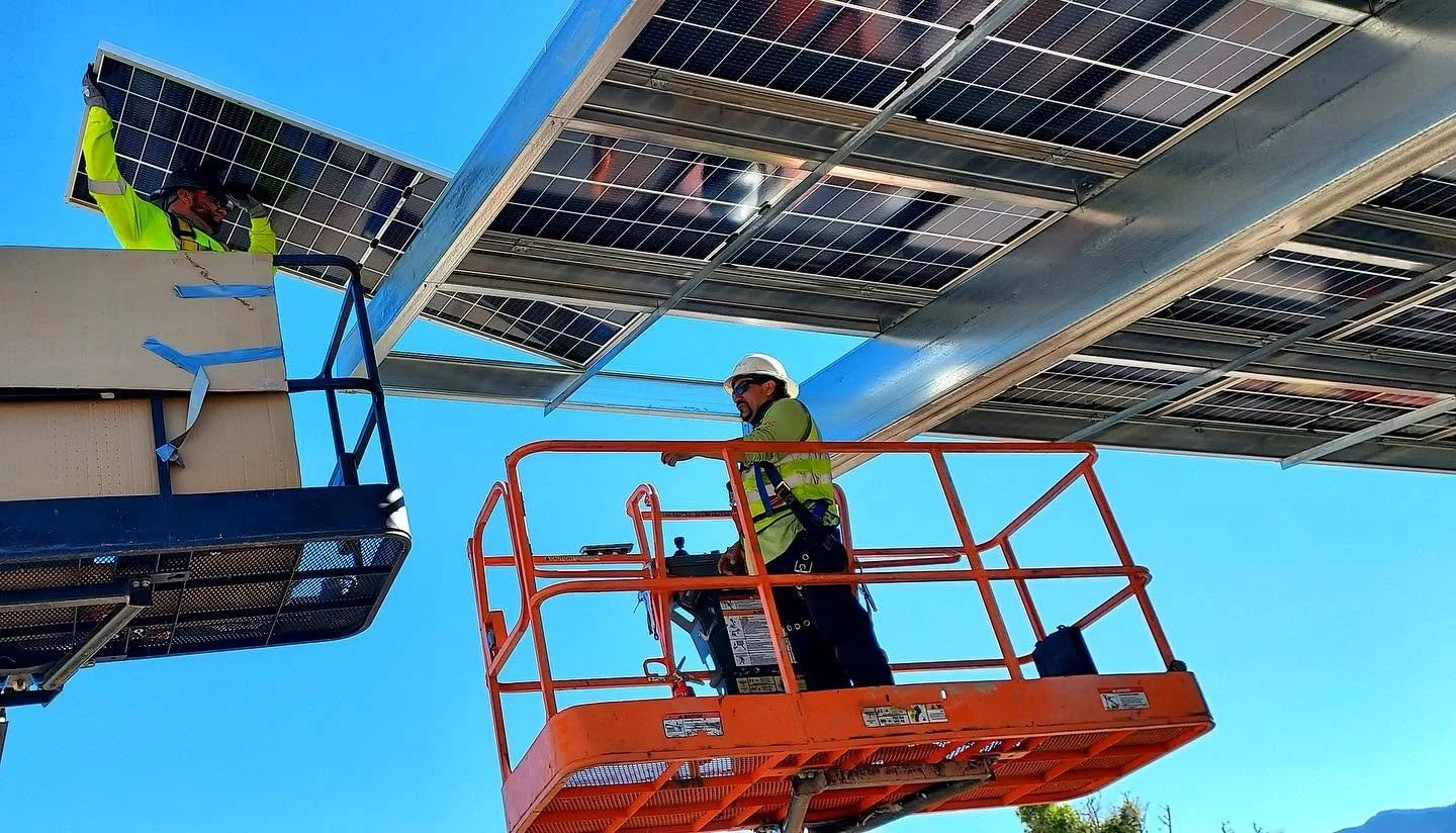 Two workers installing solar panels on a large metal structure using a lift platform, with a clear blue sky in the background.