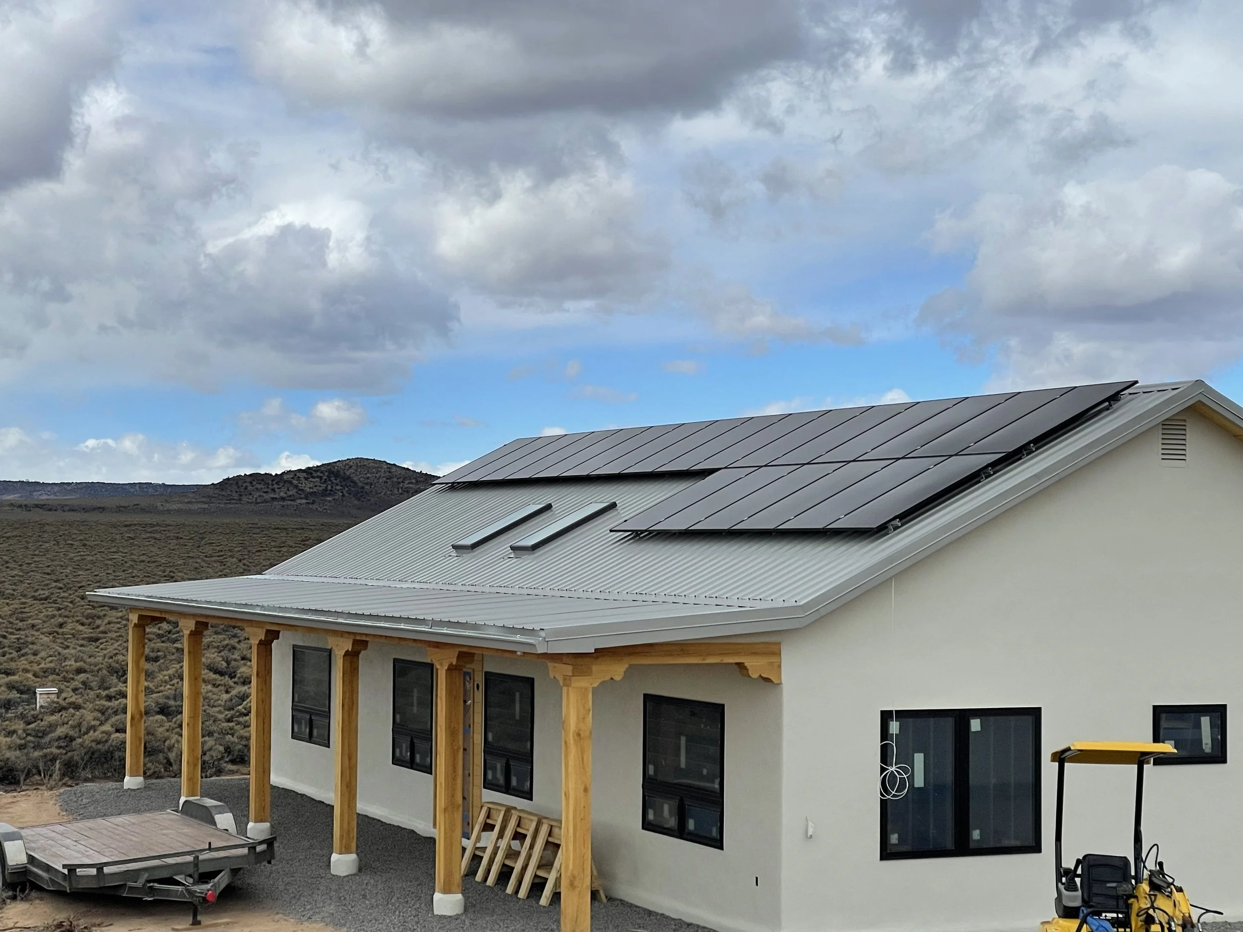 New construction home with solar panels on the roof, wooden beams supporting a porch, and large windows in a mountainous area under a partly cloudy sky.