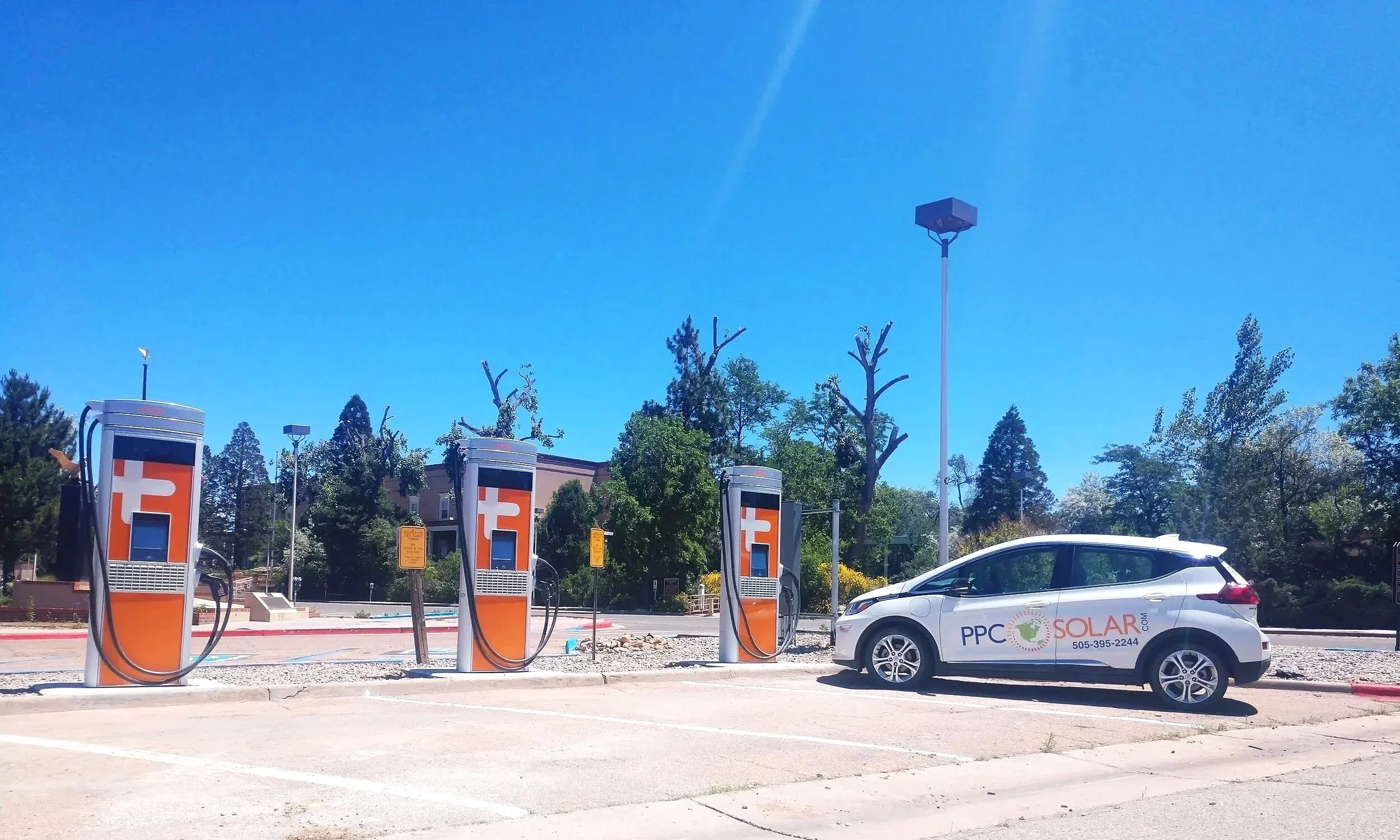 Electric vehicle charging stations with a white PPC Solar electric car parked beside them, under a clear blue sky, surrounded by trees.
