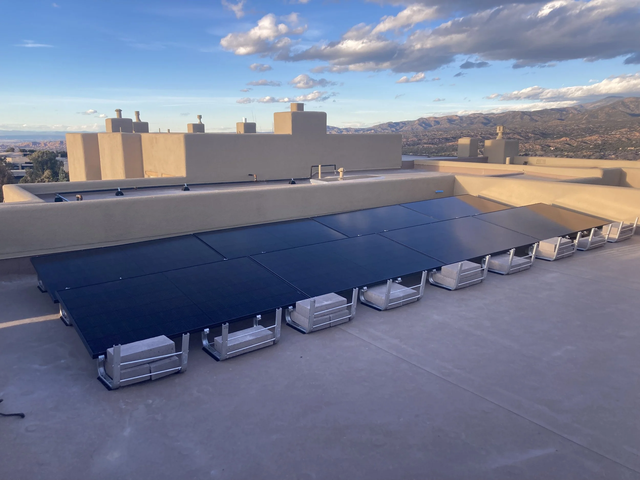 Rooftop with solar panels overlooking a city and mountain range under a partly cloudy sky.