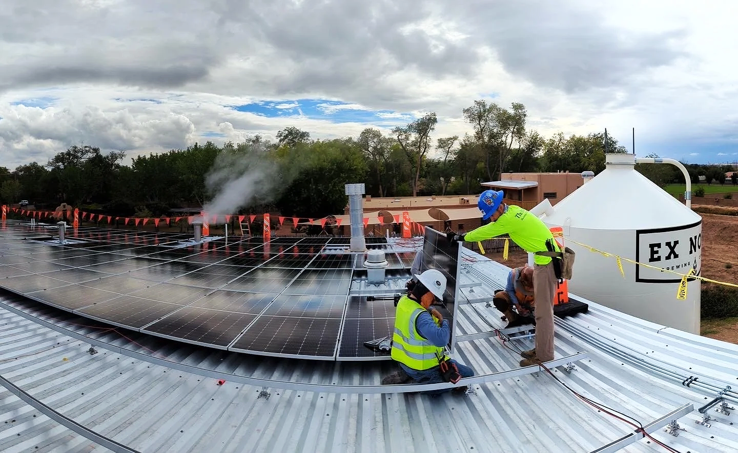 Two construction workers installing solar panels on a commercial metal roof, with a large white tank labeled 'EX NOVO Brewing' in the background.