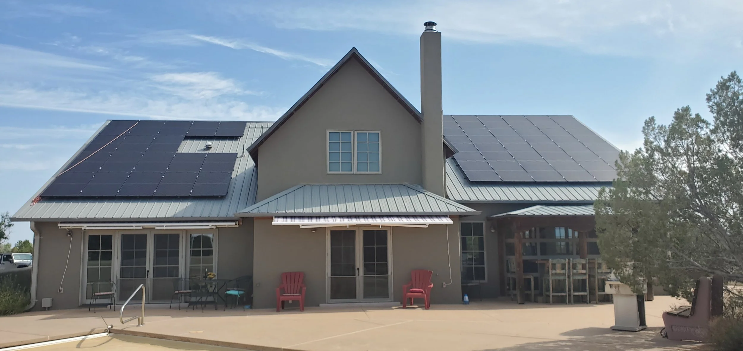 A house with solar panels on its metal roof, sliding glass doors at the back, red chairs on a concrete patio, and a tree on the right side.