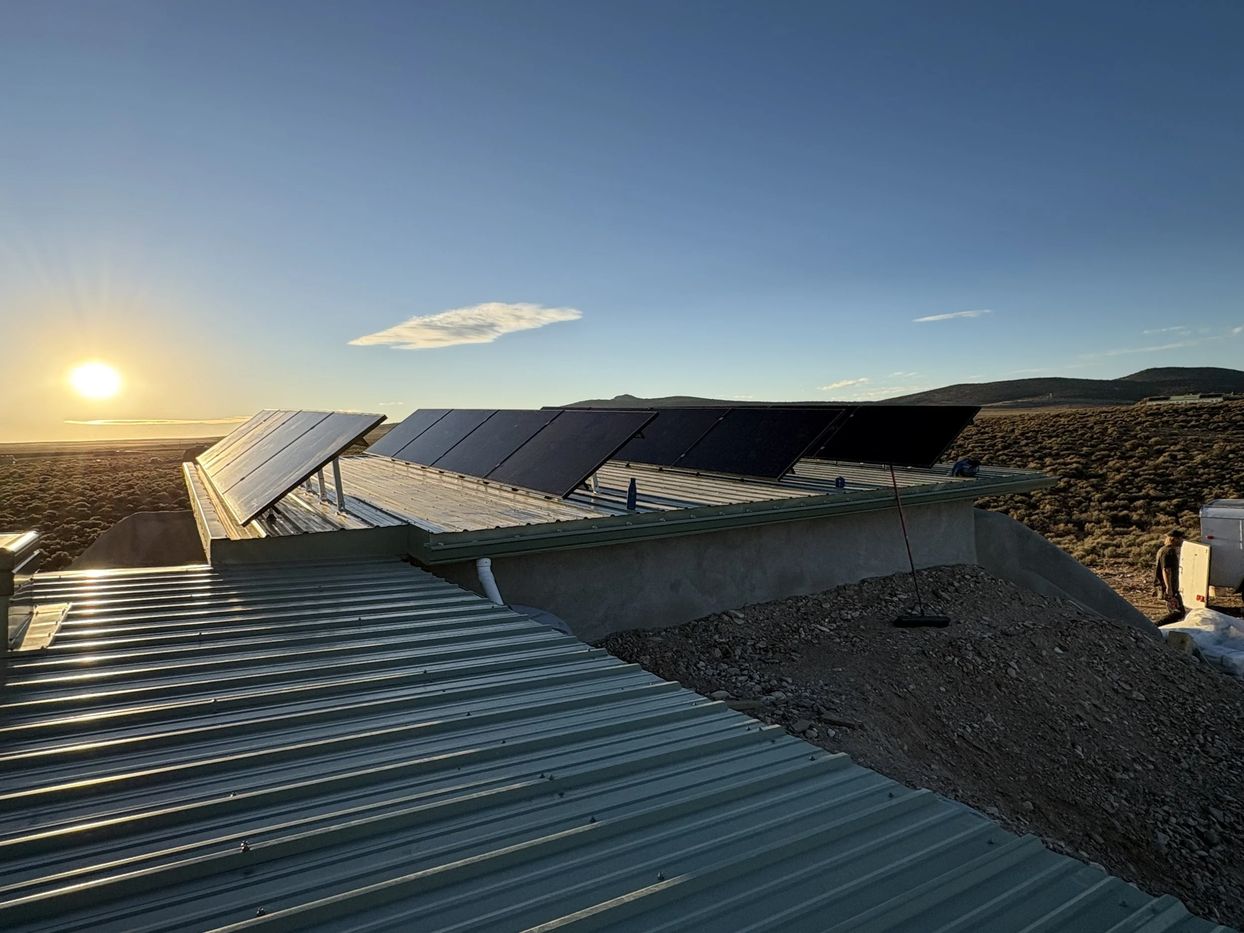 Sunset over an off grid solar panel installation on a rooftop in a rural area with hills in the background and a partly cloudy sky.