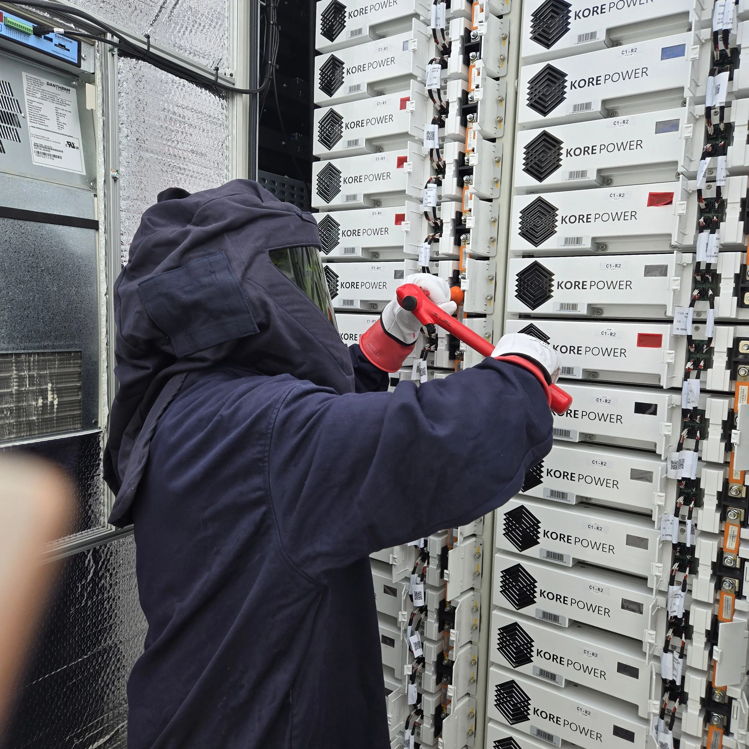 A electrical technician wearing safety gear working on a large server or electrical equipment with multiple white modules, black vents, and wiring.