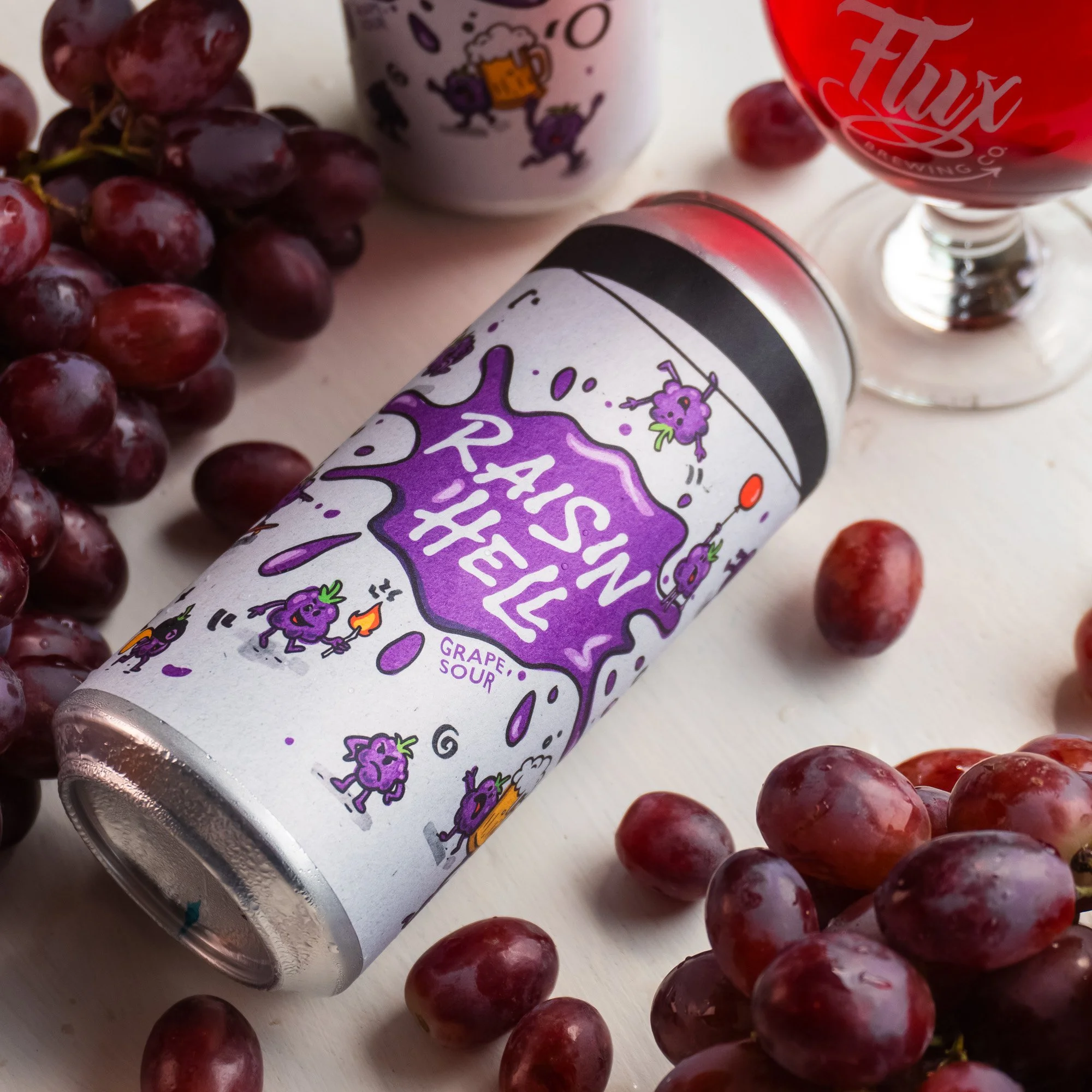 A can of grape flavoured sour beer surrounded by bunches of red grapes on a white surface.
