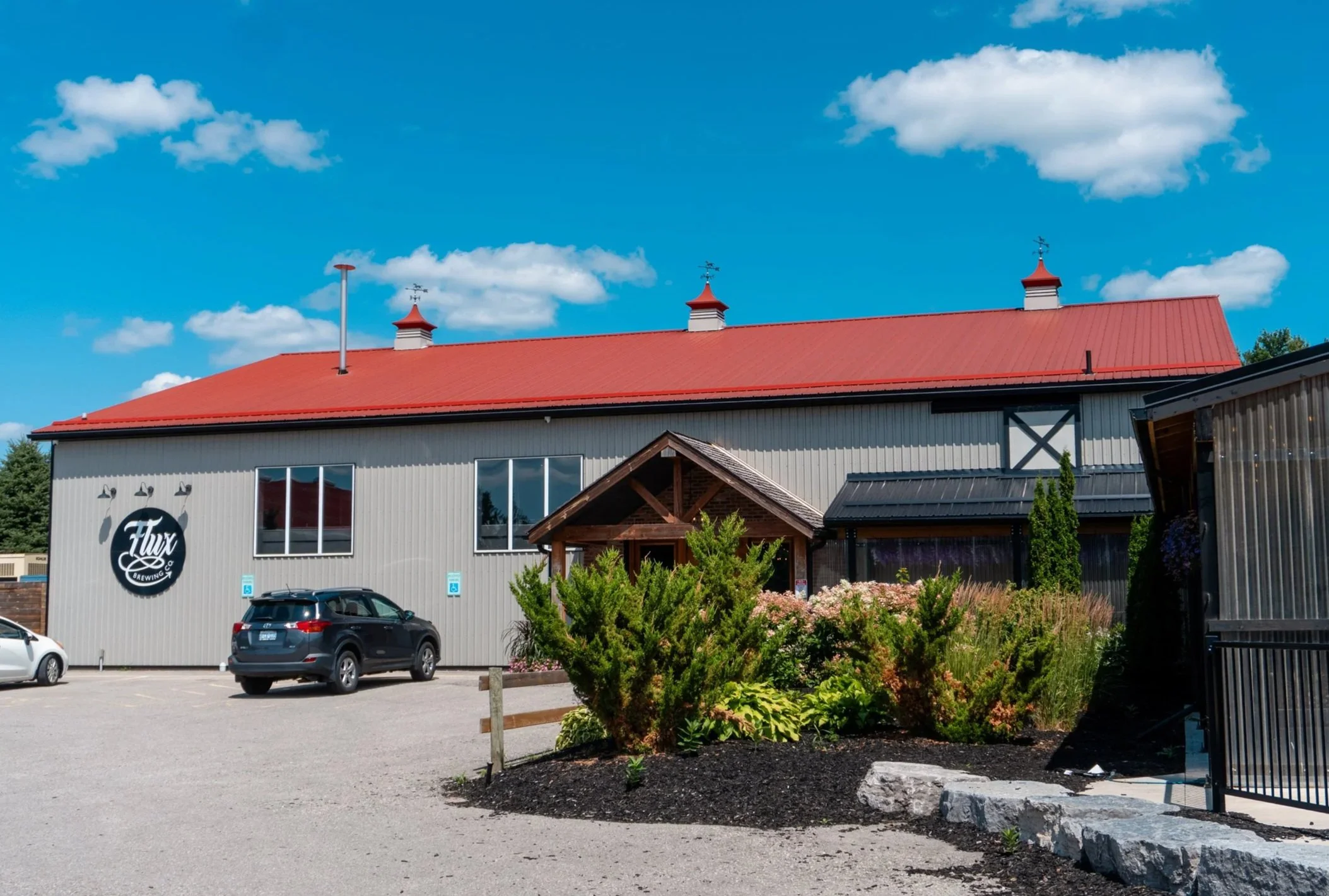 A parking lot adjacent to a brewery building with a red roof, white siding, and a circular brewery logo on the wall. There are cars parked and landscaped bushes and flowers in the foreground.