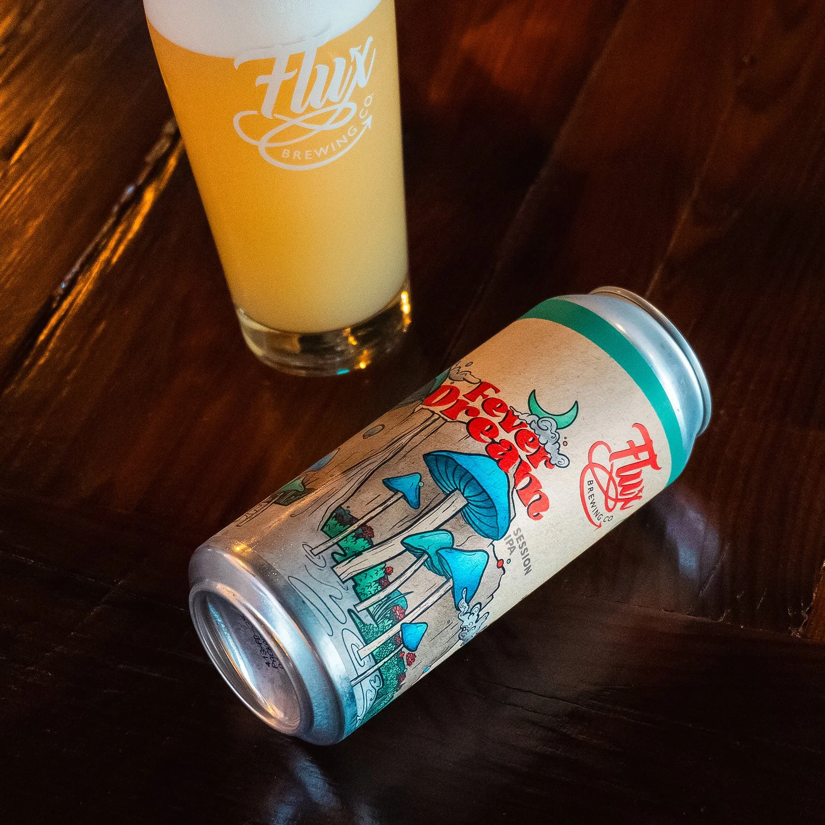A can of beer with colorful mushrooms and waterfalls on the label, placed on a blue background with outlined mushroom illustrations, alongside a glass of beer.