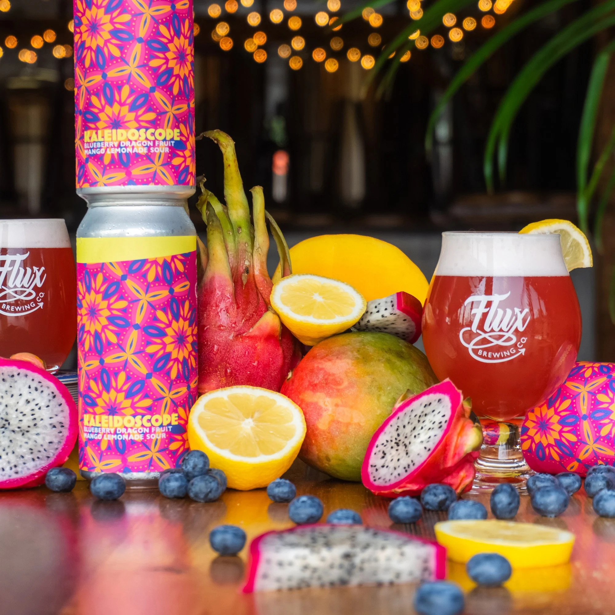 Colourful beer cans alongside fresh fruits, including lemons, blueberries, dragon fruit, mango, and lemon slices, on a wooden surface with blurred warm string lights in the background.