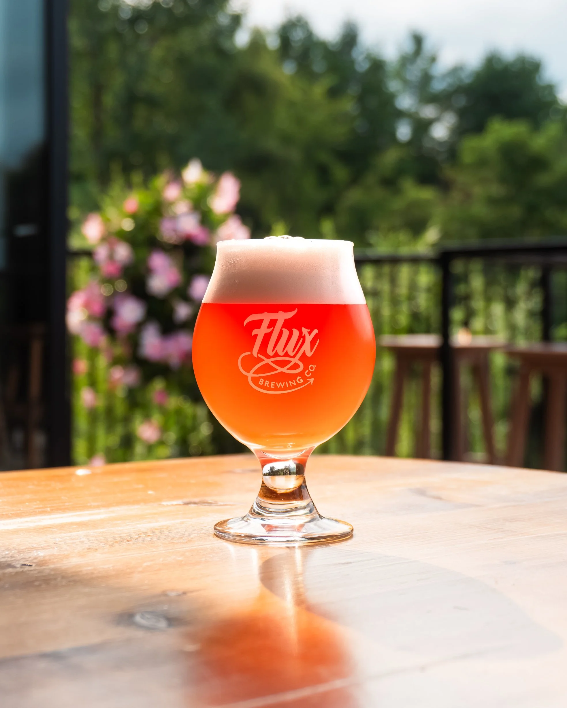 A glass of pinkish orange beer with frothy head on a wooden table outdoors, blurred green trees and pink flowers in the background.