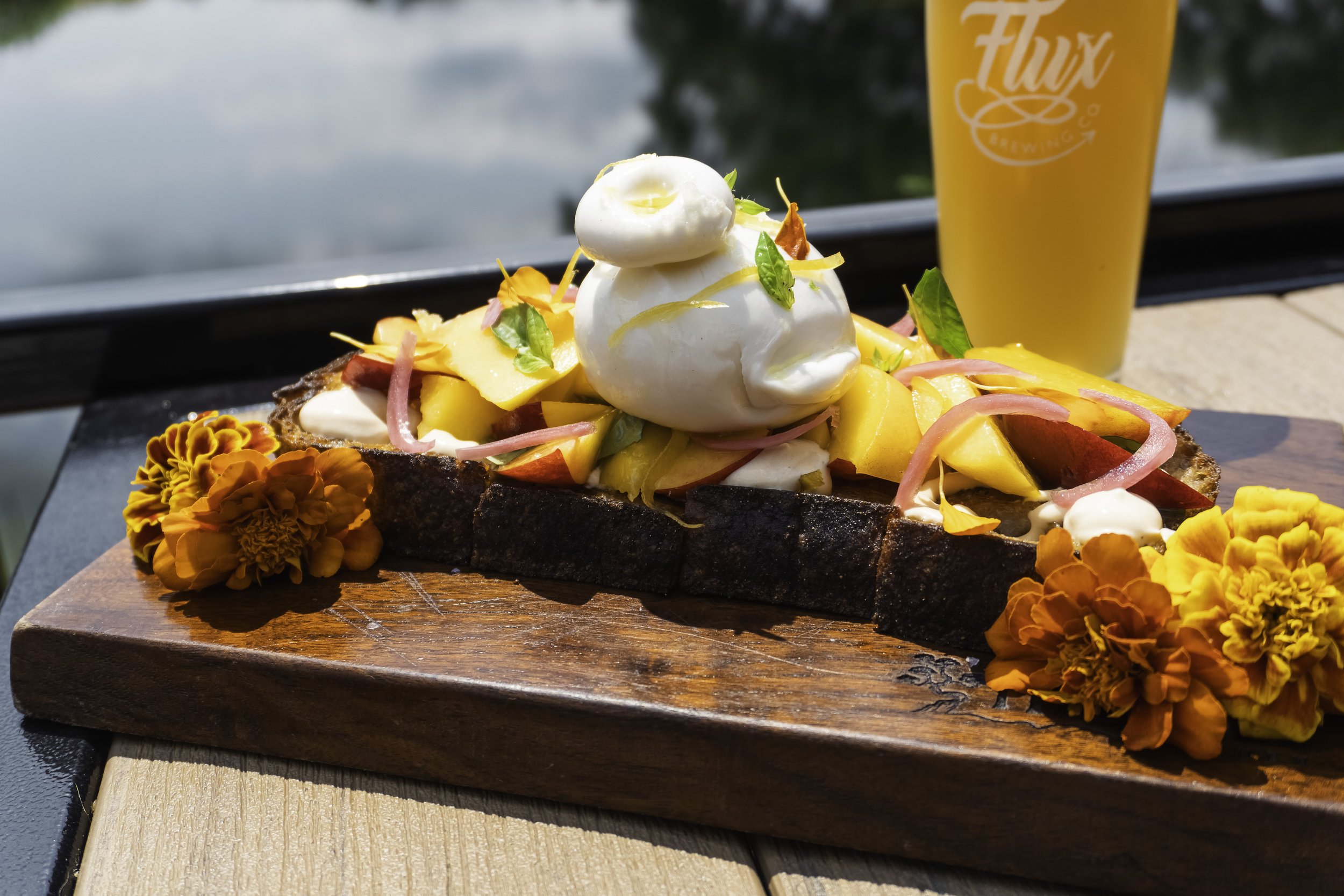 A piece of toasted bread topped with sliced peaches and burrata cheese, with a hazy beer and pond in the background.