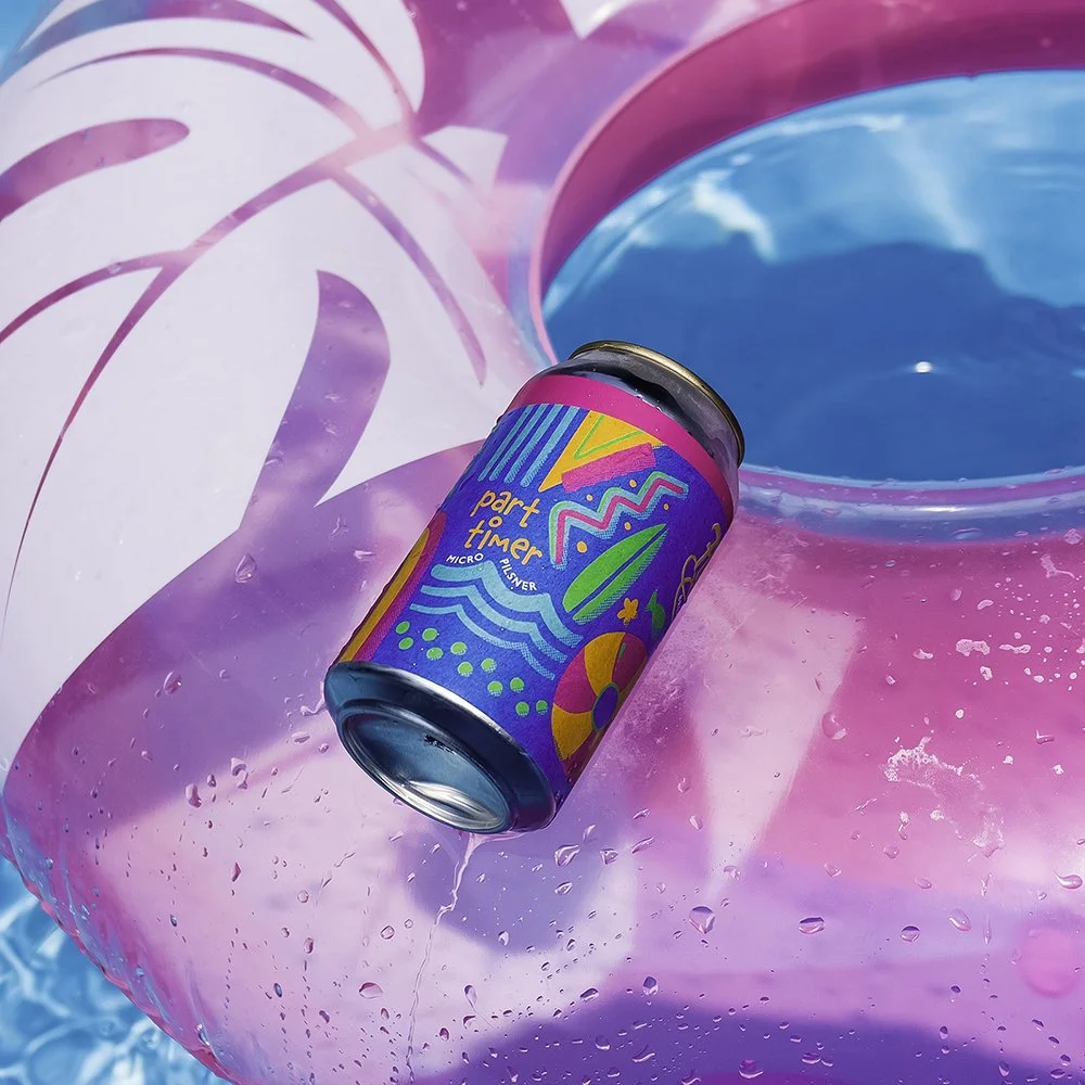 A colorful can with vibrant abstract designs and the words 'part timer' floating on a pink and purple inflatable pool float with water droplets, in a pool.