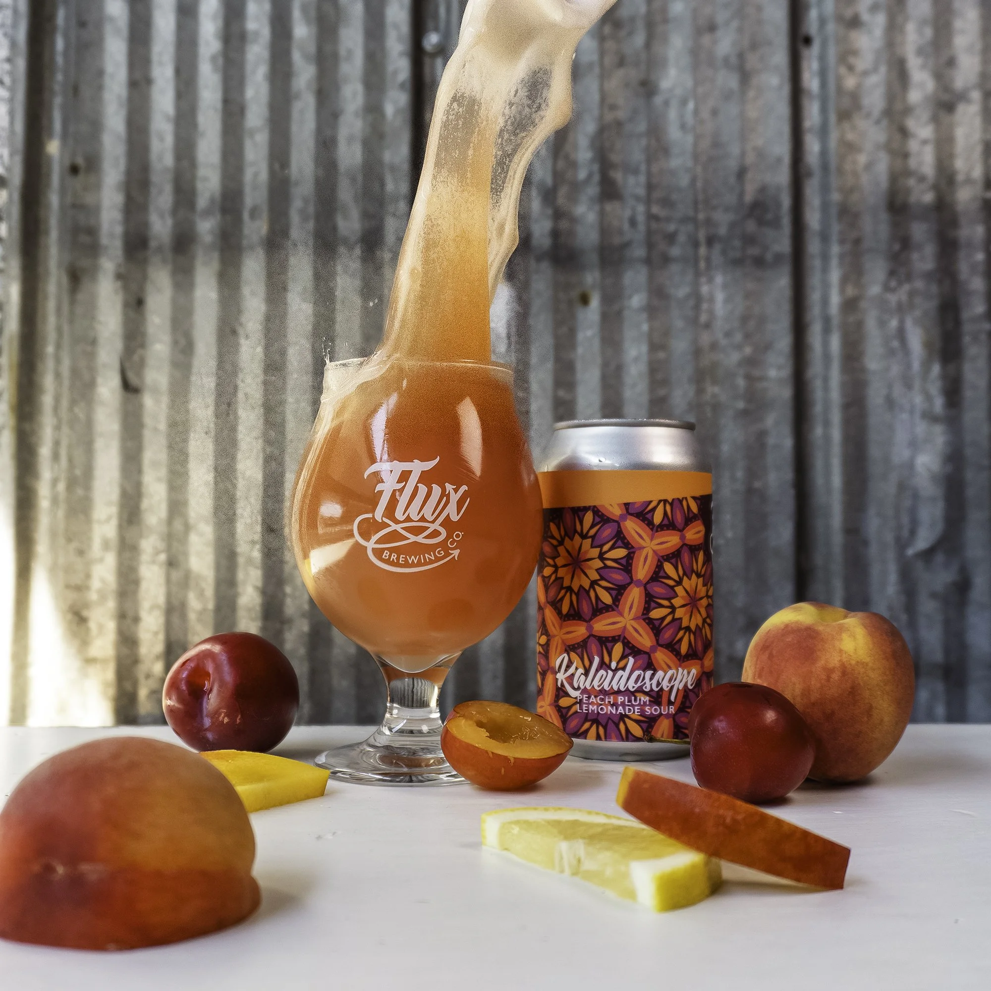 A glass of peach plum lemonade sour beer with beer splashing out of the glass, surrounded by peaches, plums, lemon slices, and a colourful can of Kaleidoscope beer on a white surface with a wooden background.