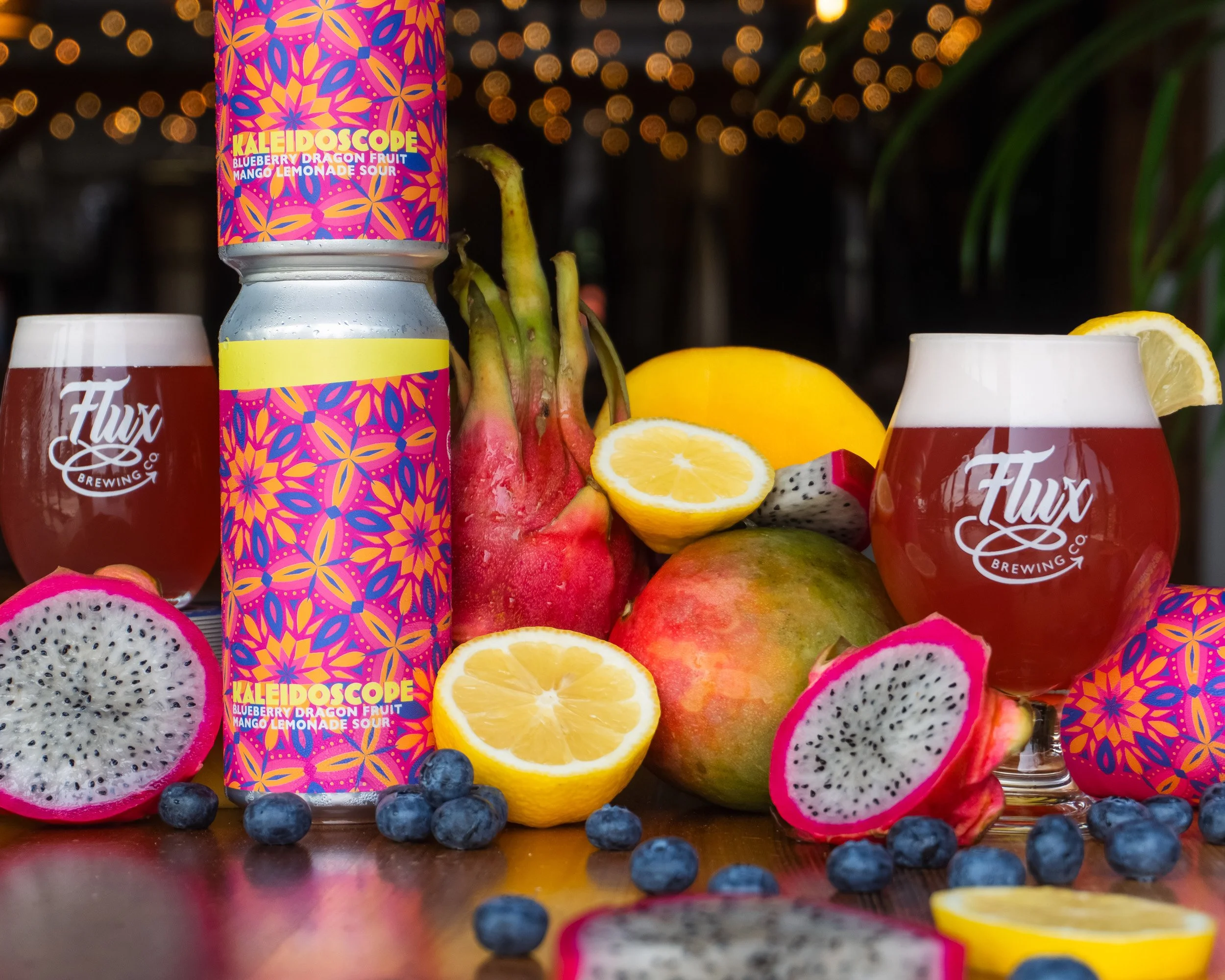 Close-up of a colorful can of Kaleidoscope sour drink with blue berries, sliced lemon, dragon fruit, and other fruits around it, against a background of string lights.