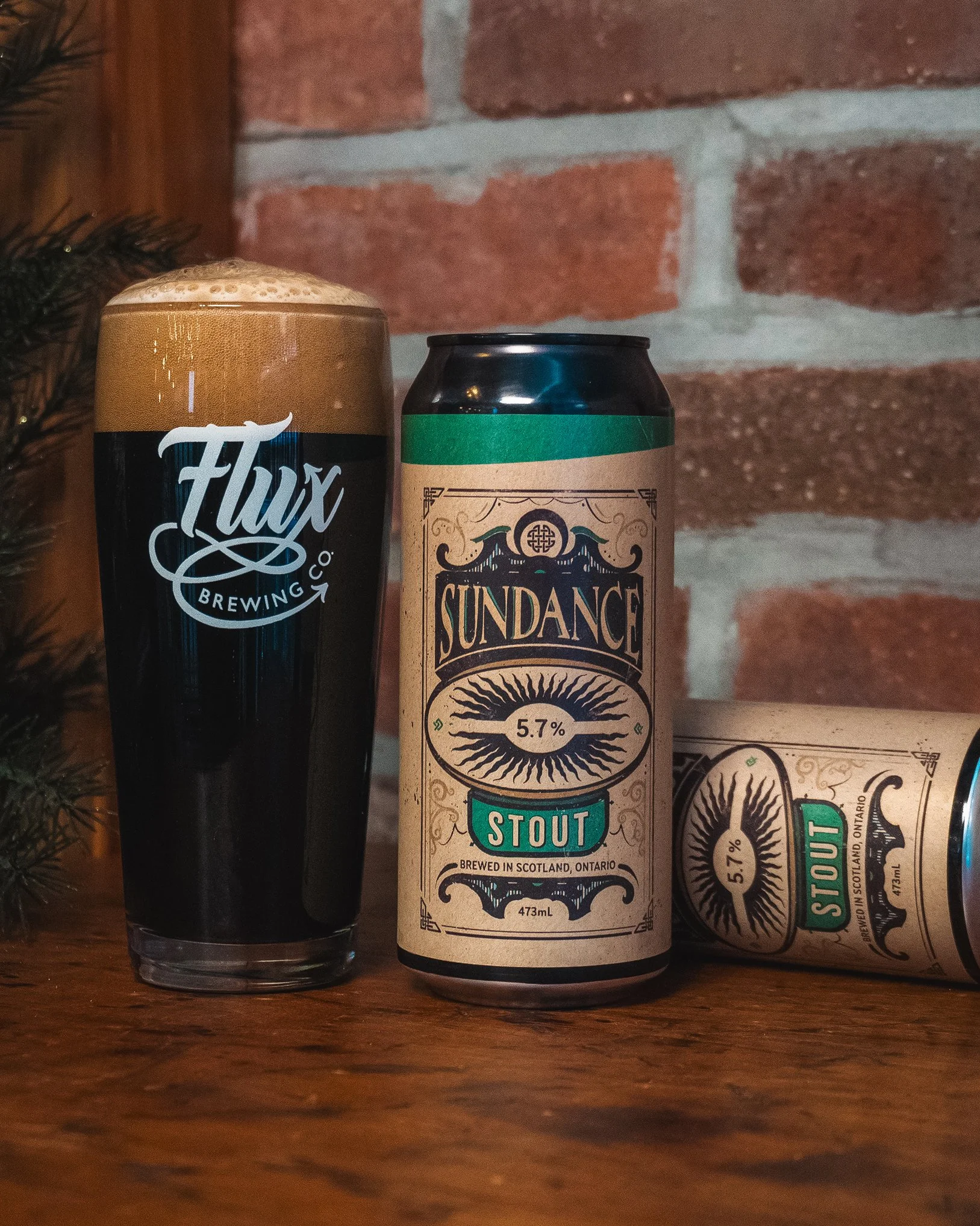 A glass of dark stout beer with a frothy head from Flux Brewing Co. and a can of Sundance Stout, brewed in Scotland, Ontario, on a wooden surface against a brick wall background.