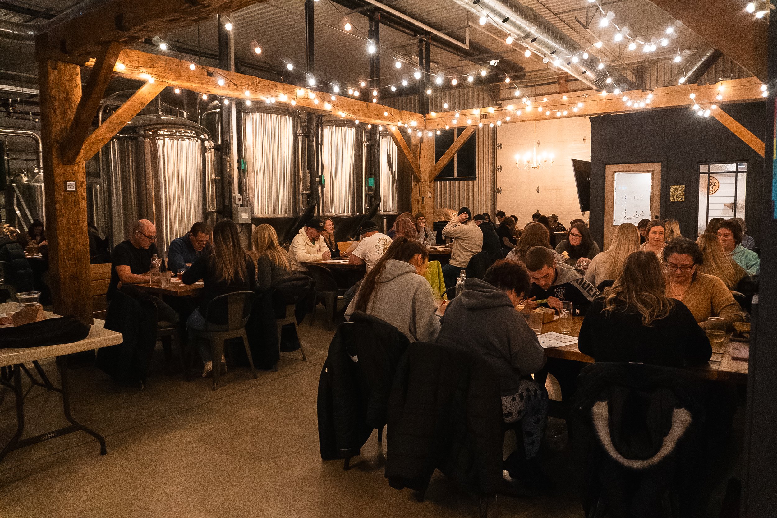 People dining inside a well-lit brewery with large brewing tanks in the background and string lights overhead.