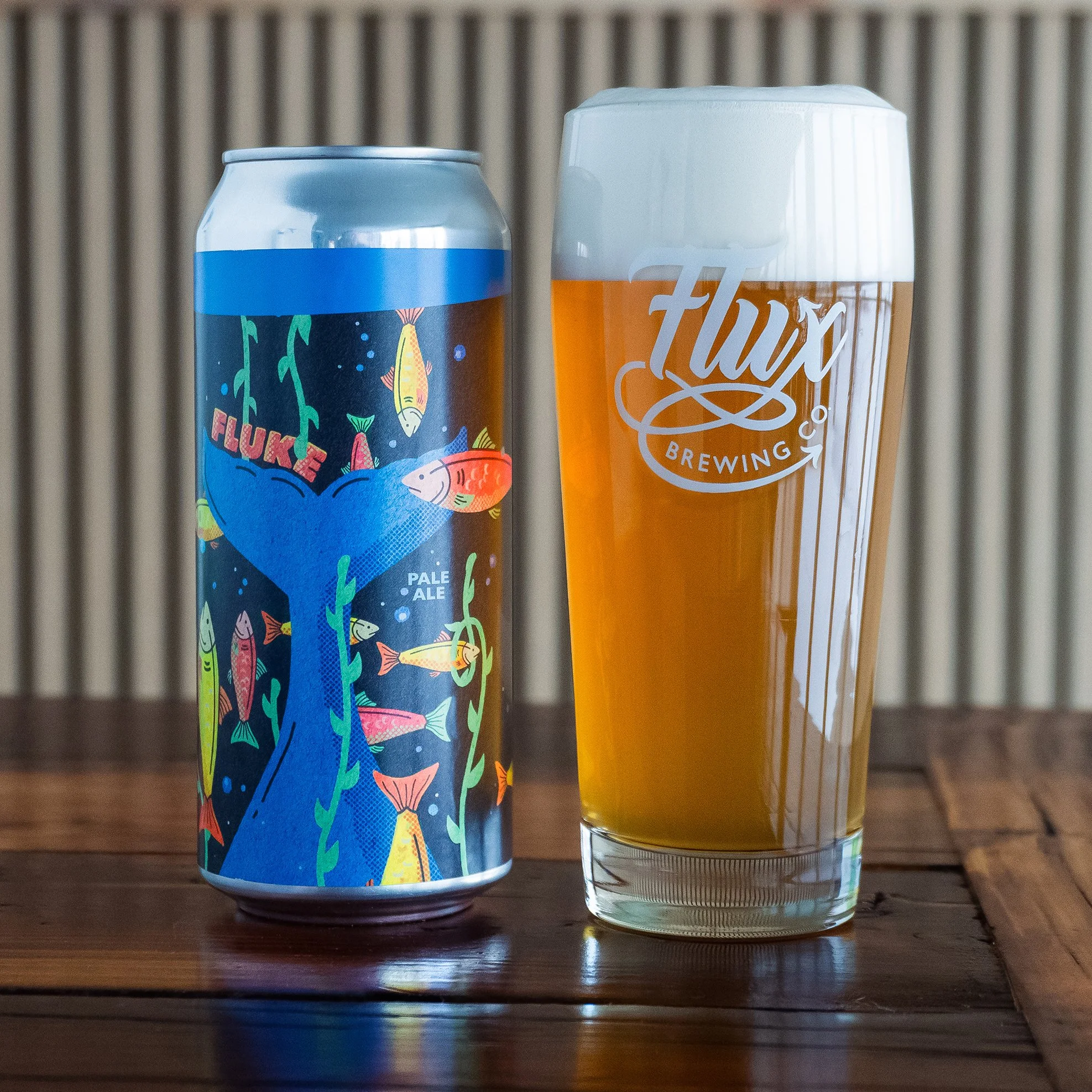 A can of Fluke Pale Ale with colorful fish and whale tail illustrations next to a Flux Brewing Co. pint glass filled with beer on a wooden surface.