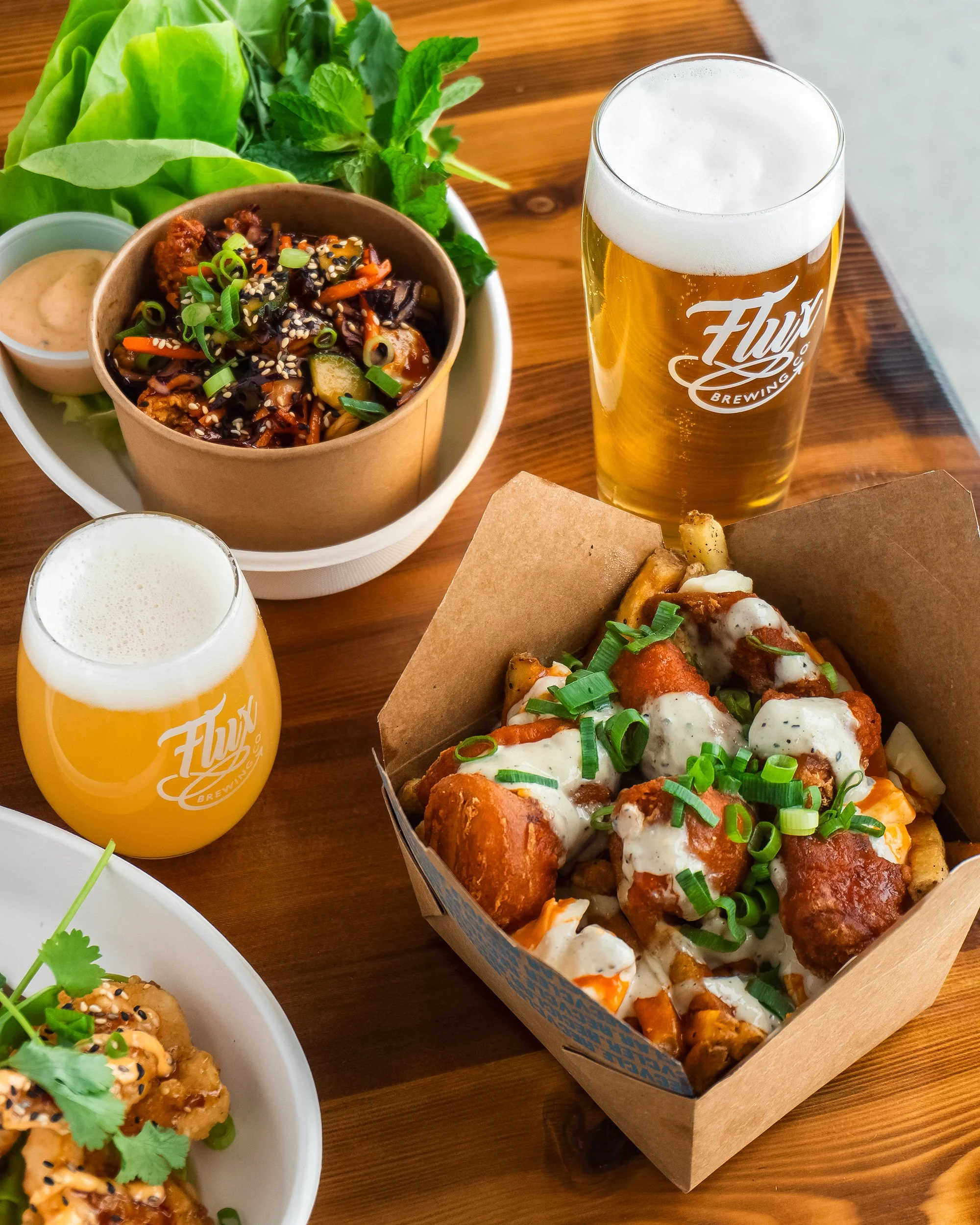 Two glasses of beer, a box of loaded buffalo fries, a bowl of Asian-inspired stir-fry lettuce wraps on a wooden table.