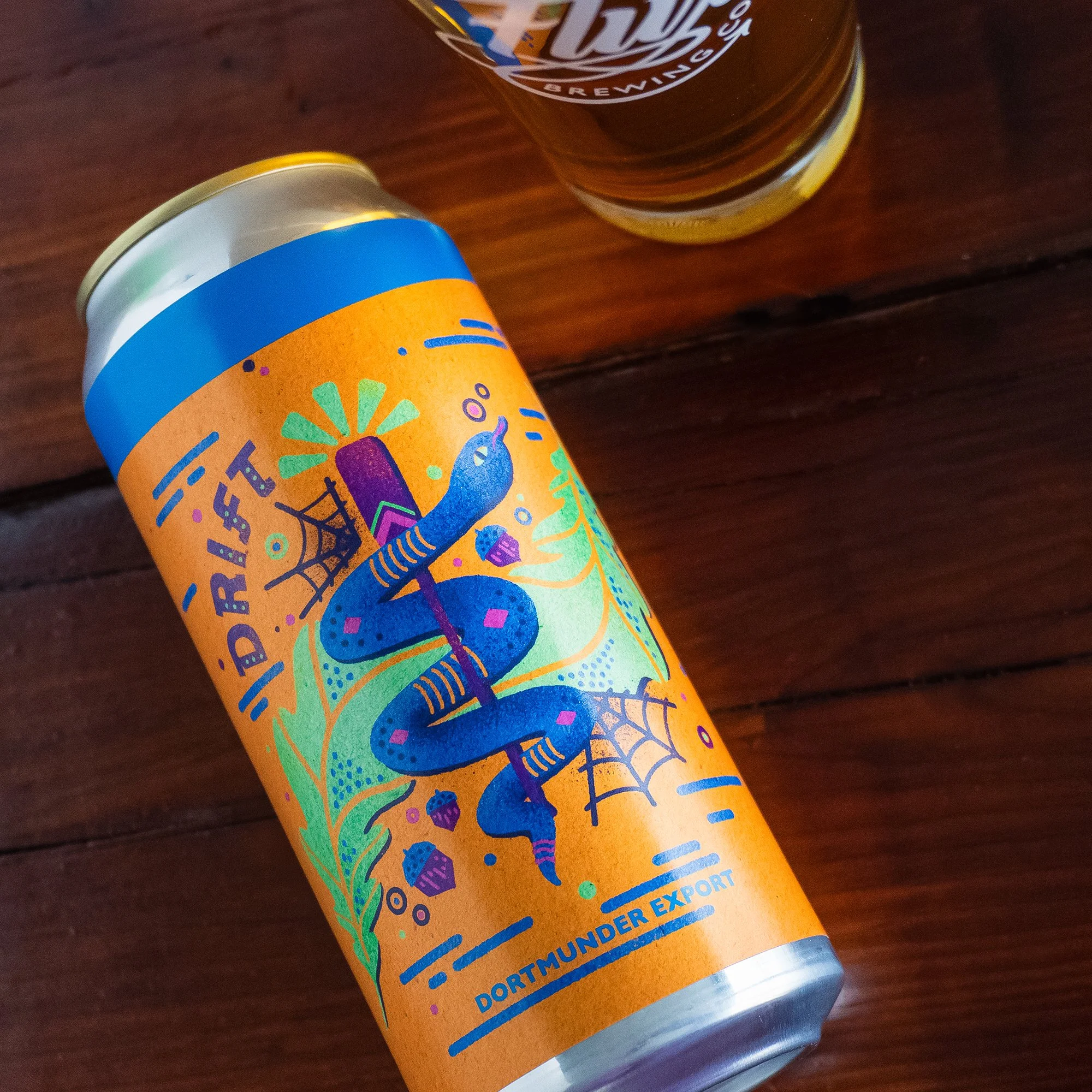 A can of Dortmunder Export beer with a vibrant label featuring a blue snake wrapped around a purple oar, spiderwebs, acorns, and the word 'Drift' on an orange background, placed on a dark wooden surface with a glass of beer partially visible.