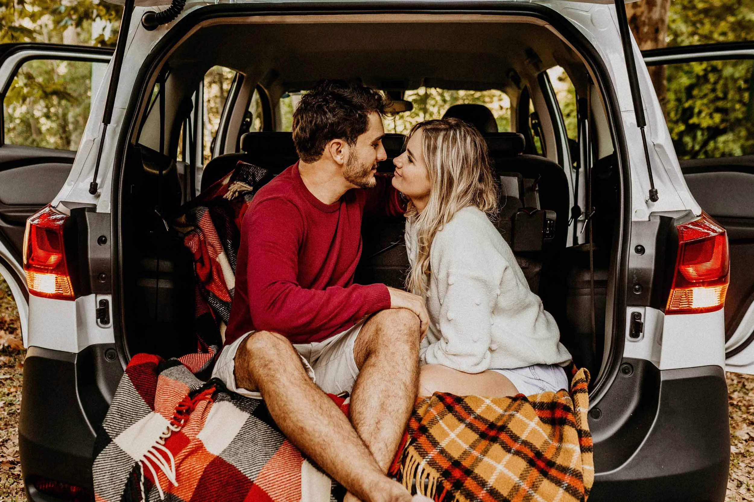 A couple sitting closely in the back of an SUV, leaning towards each other with their faces close, surrounded by fall foliage, on a plaid blanket.