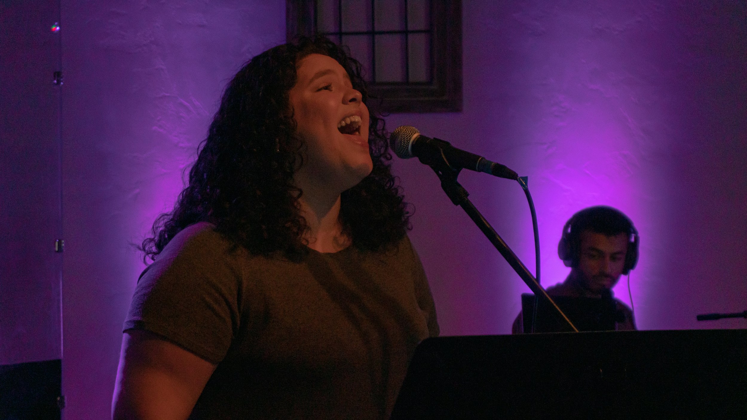 A woman with curly hair singing into a microphone, with a person in the background wearing headphones, in a dimly lit room with purple lighting.