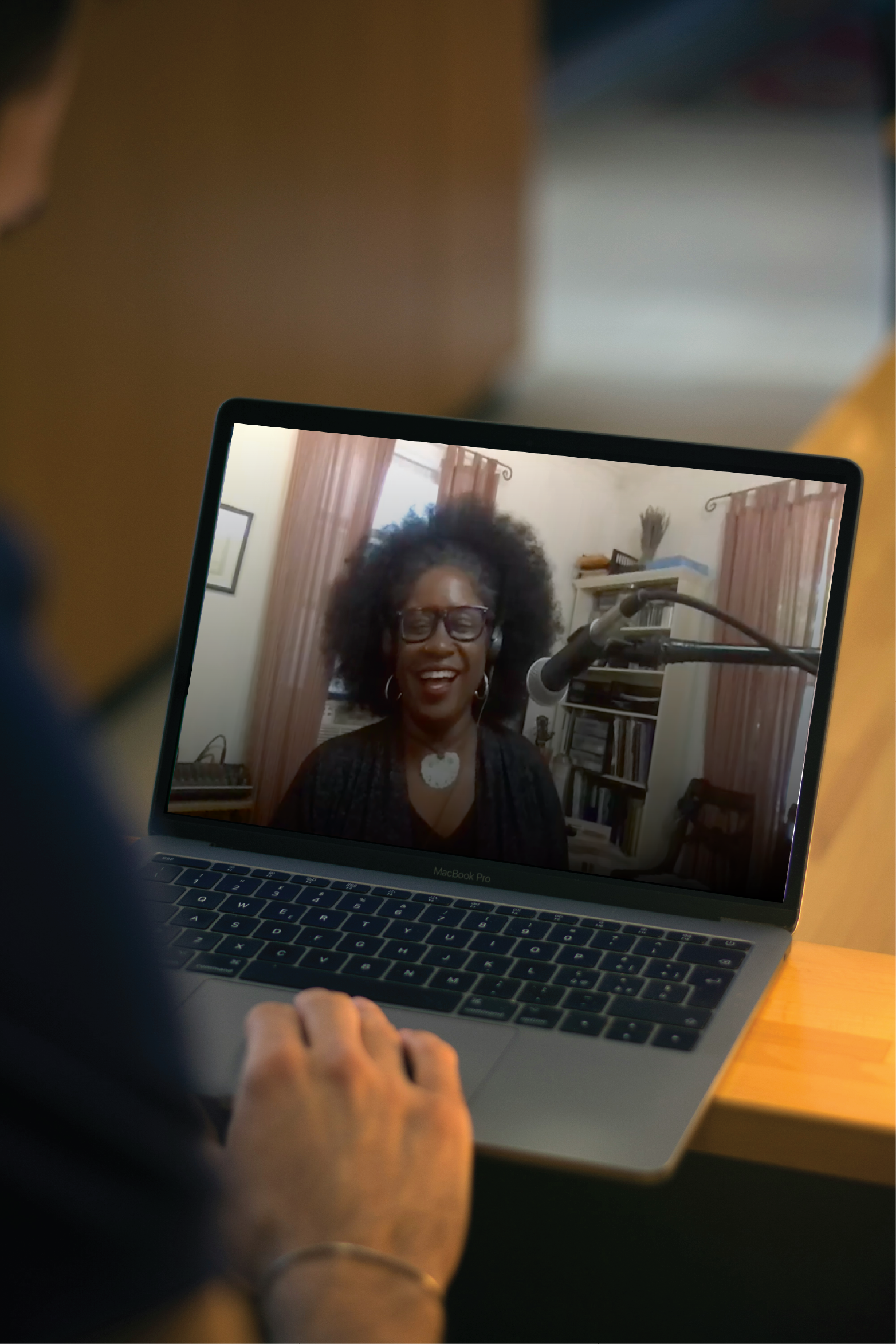 A person using a laptop shows a video call with a woman wearing glasses, a black top, and a necklace, smiling with curly hair in a room with bookshelves, curtains, and a window.