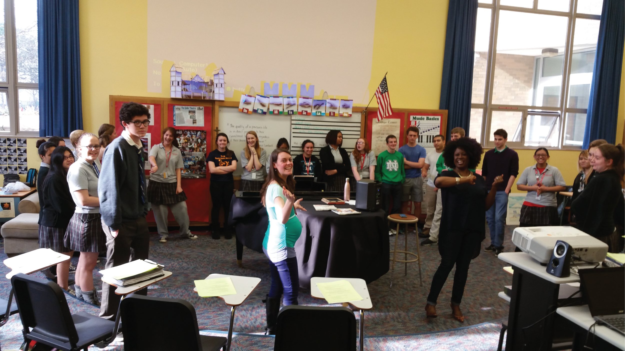 Group of students and teacher gathered in a classroom, some smiling and talking, with posters and windows in the background.