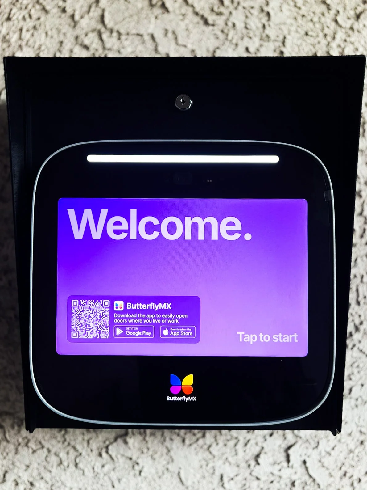 A ButterflyMX smart door intercom device displaying a purple welcome screen with a QR code, app download prompts, and the text 'Tap to start'.