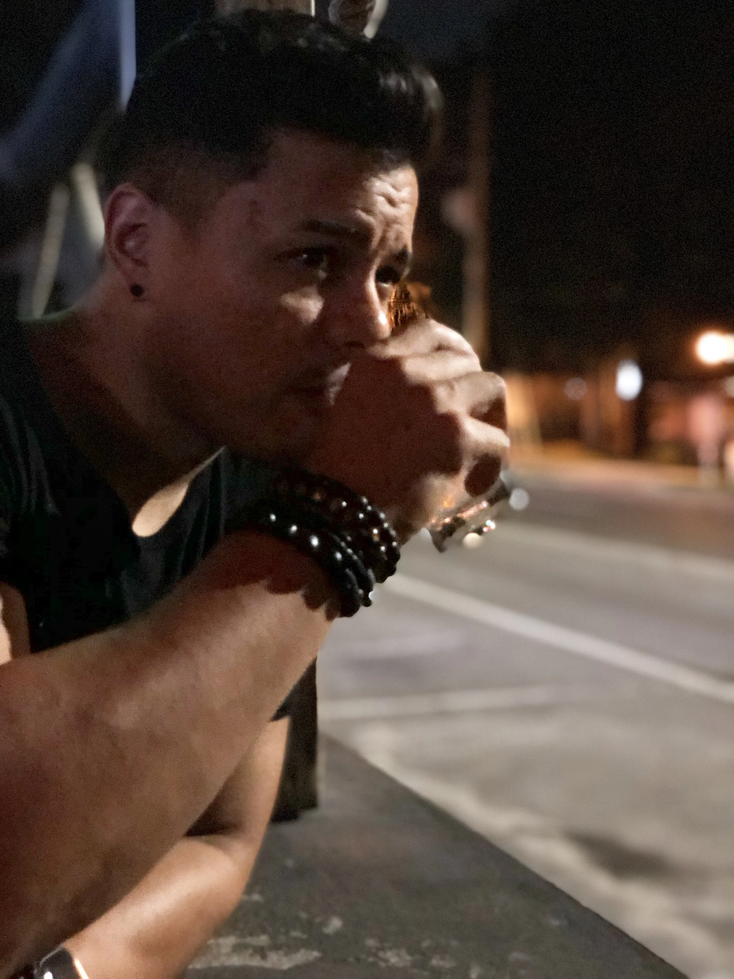 A person with short, dark hair and earrings leans against an outdoor bar top at night, having a beverage. They are wearing black clothing with a bracelet on their wrist. The background shows a street with blurred lights.
