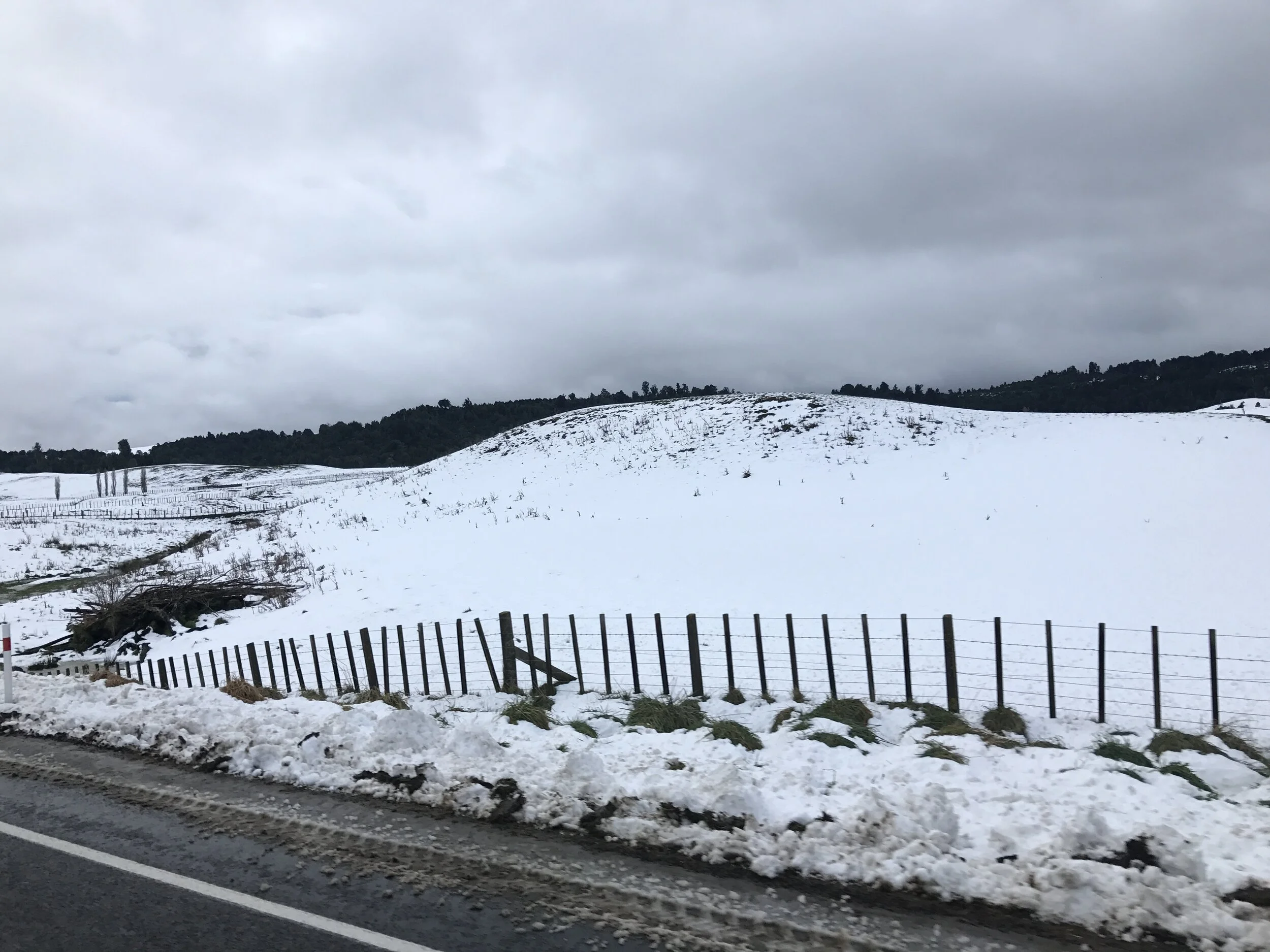 Snow-covered landscape seen from a roadside, with a dark fence in the foreground, rolling hills and trees in the distance, and a cloudy sky overhead.