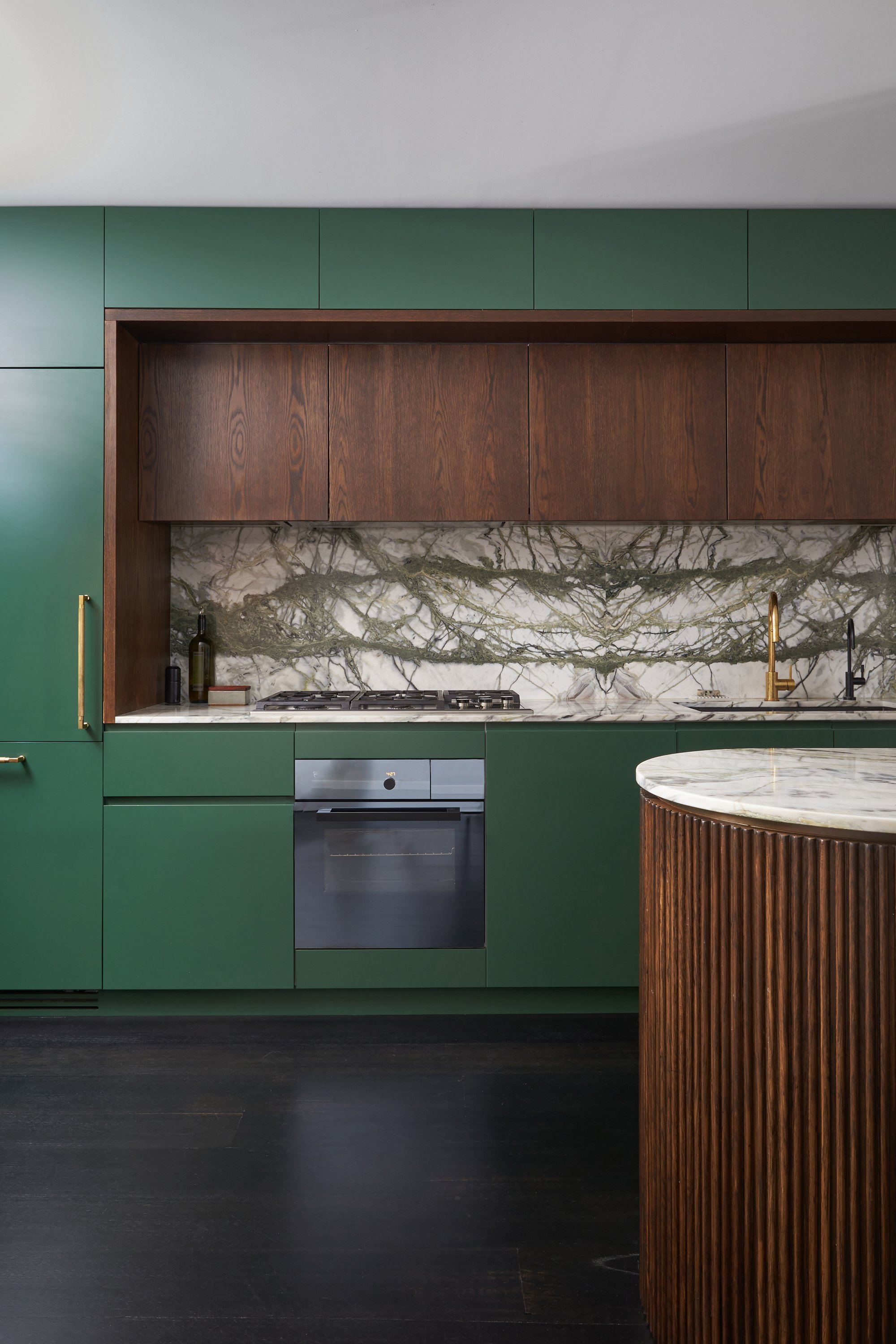 Modern kitchen with black cabinetry and white countertops, large floor-to-ceiling windows with city and greenery views, a vase with purple flowers on the island, and minimal decorative items.