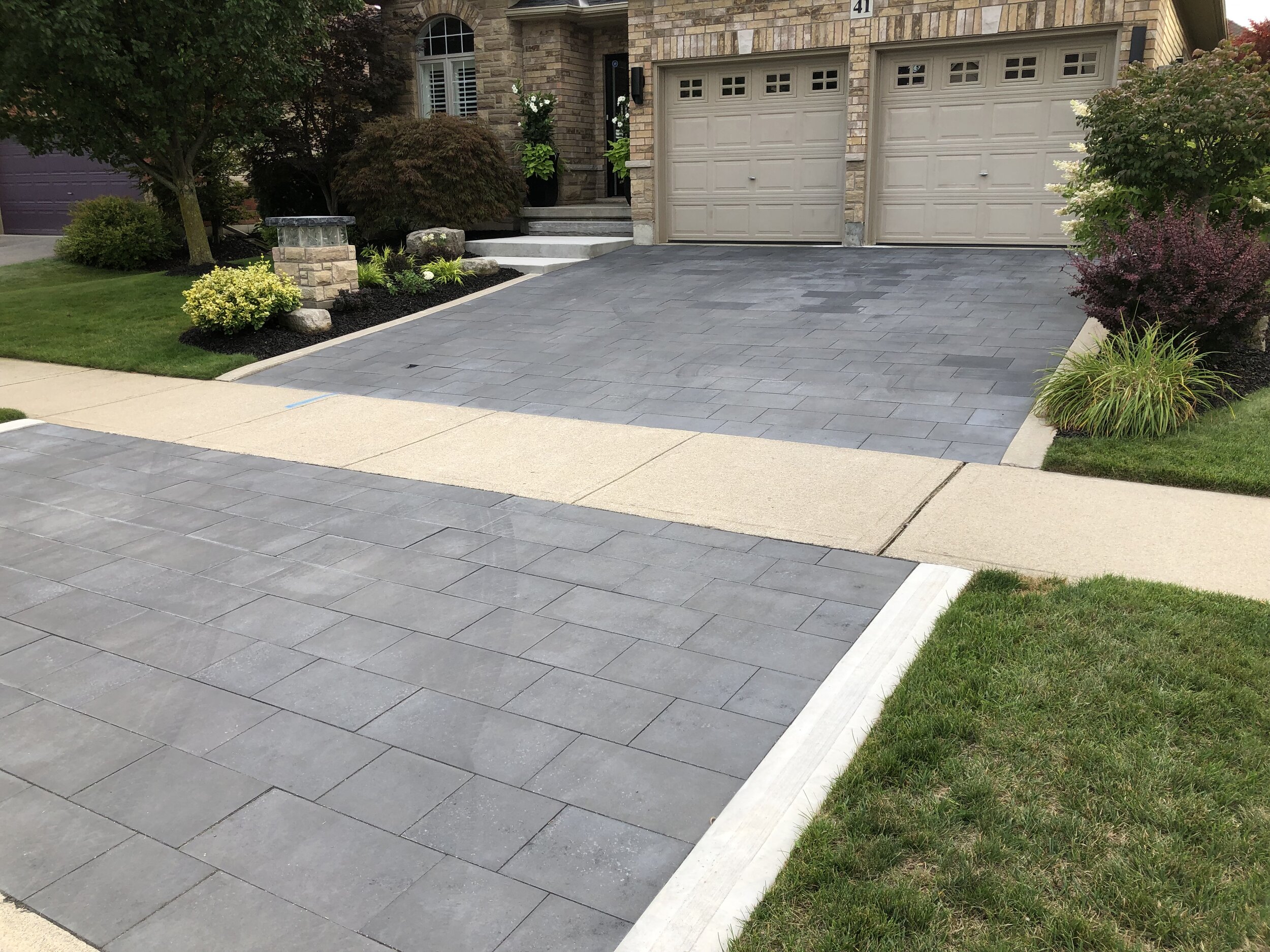 Newly paved driveway with gray stone tiles, adjacent to a well-manicured lawn and garden. Two garage doors are visible in the background.