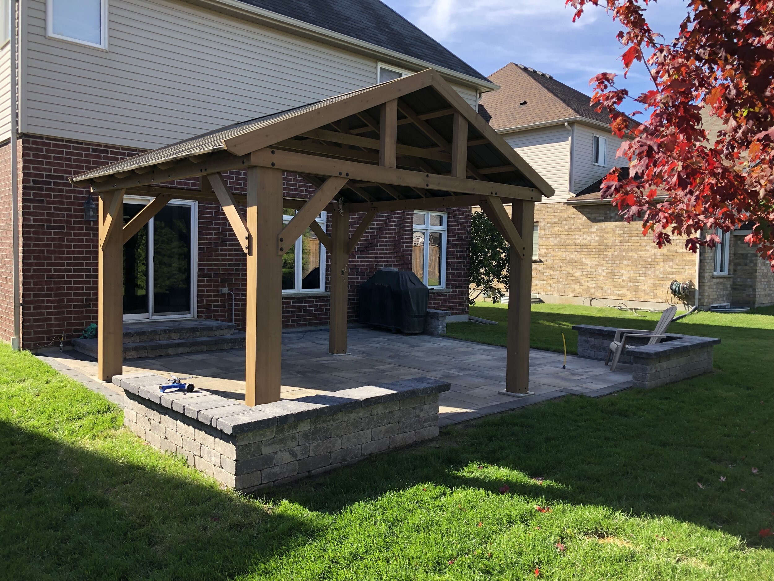 Backyard patio with wooden pergola structure and patio furniture, adjacent to a brick and siding house, with lawn and a red-leaf tree nearby.