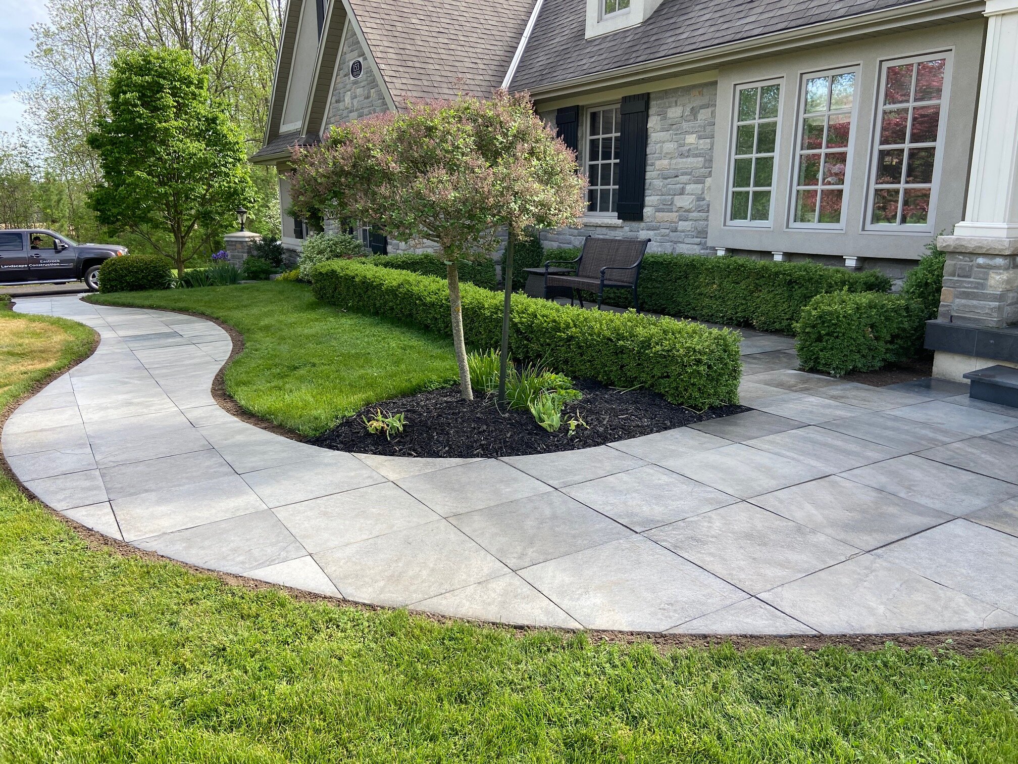 A curved stone pathway leading to the front entrance of a house with well-maintained landscaping, including a tree, shrubs, and green grass. The house features a stone facade, white-framed windows, and a sloped roof. A truck is parked on the driveway