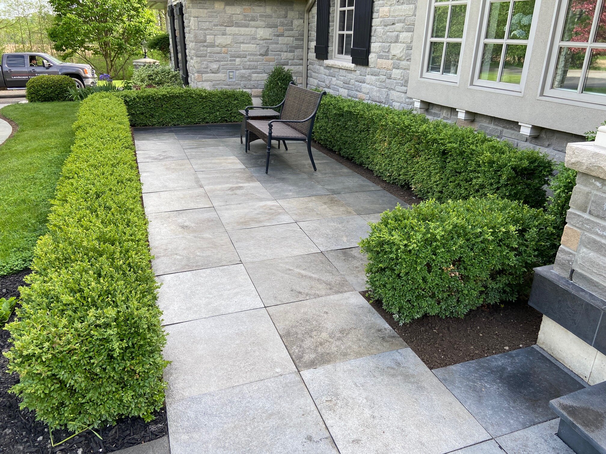 Stone patio with two chairs, surrounded by manicured bushes and a stone house exterior.
