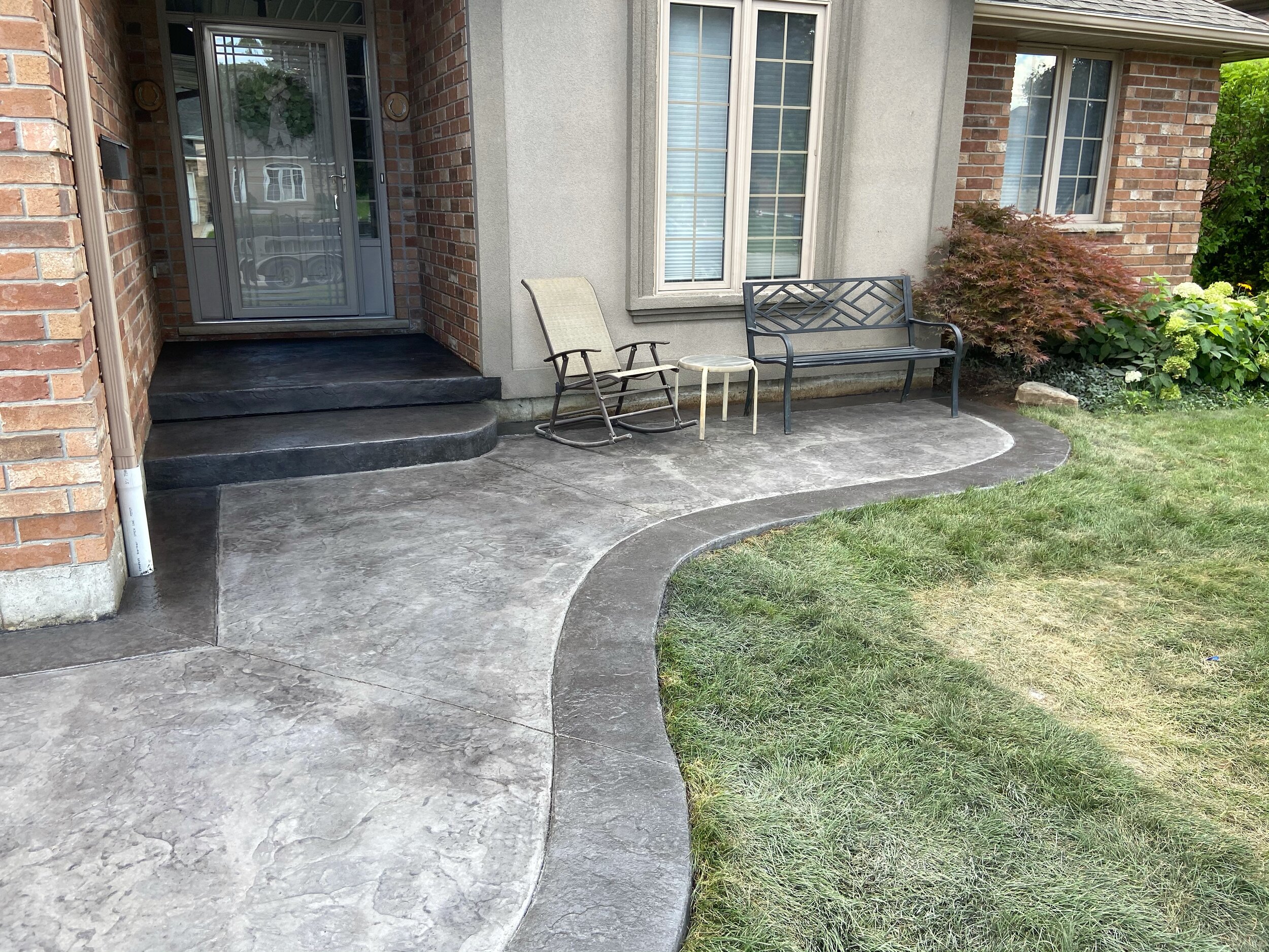 Front porch with stamped concrete walkway, a bench, a rocking chair, and shrubs.