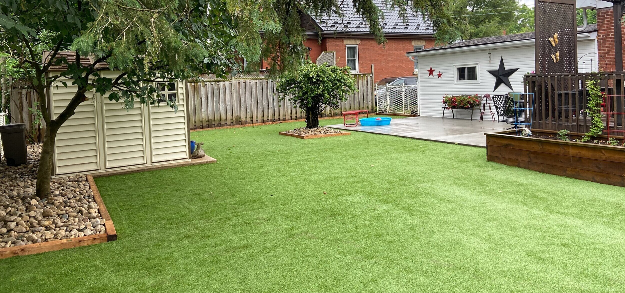 Backyard with artificial lawn, garden shed, tree, patio with potted plants, and decorative stars on the house exterior.
