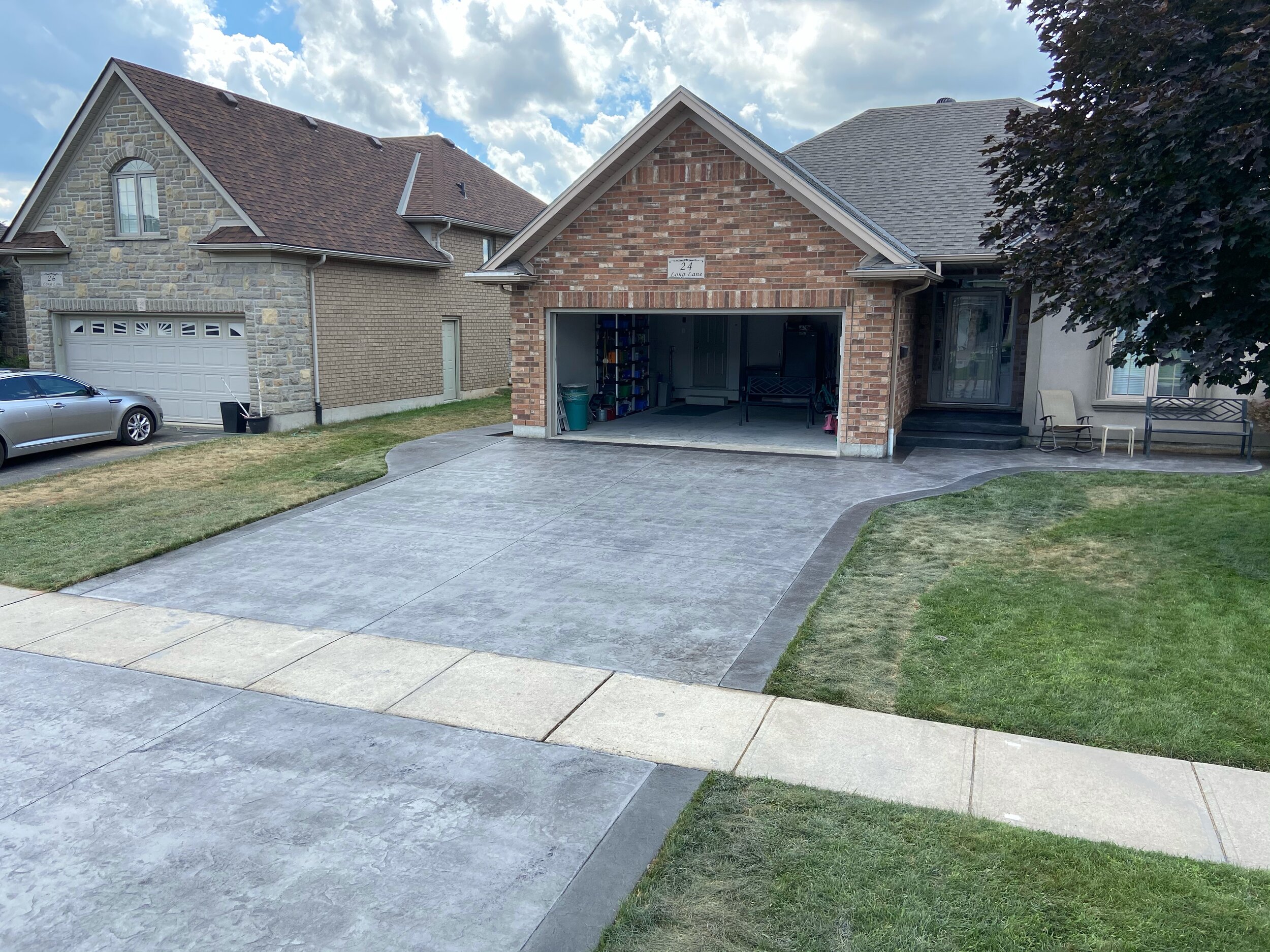 Residential scene with a brick house, open garage, paved driveway, adjacent house with stone facade, and a parked sedan. A well-maintained lawn and a sidewalk are visible in the foreground.