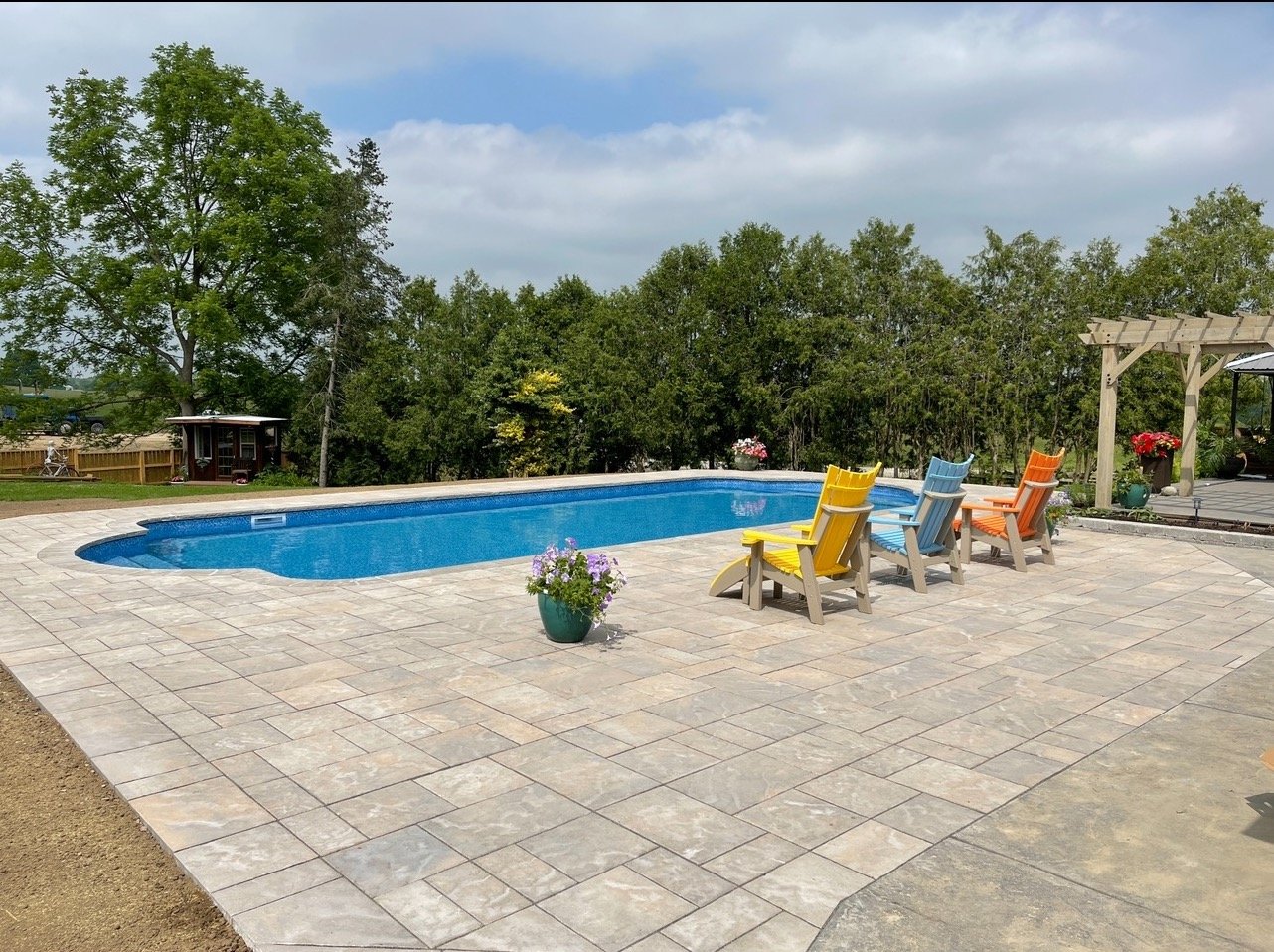 Backyard swimming pool with colorful chairs, paved patio, and surrounding trees.