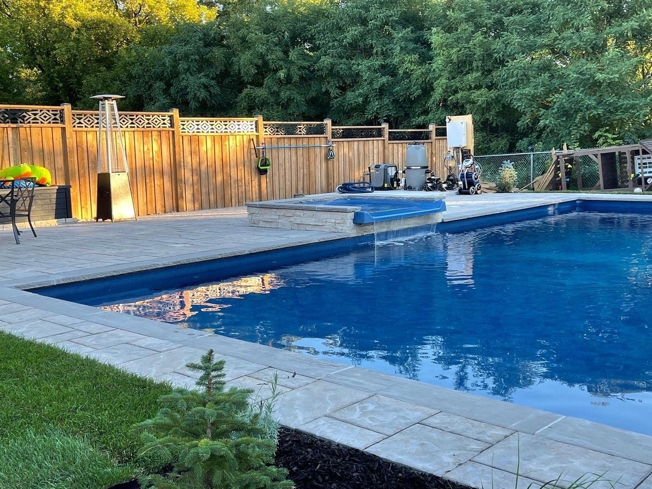 Backyard swimming pool area with patio, trees, and a wooden fence. Pool equipment is visible near the fence. A tall heater and a small table with a colorful item are on the deck.