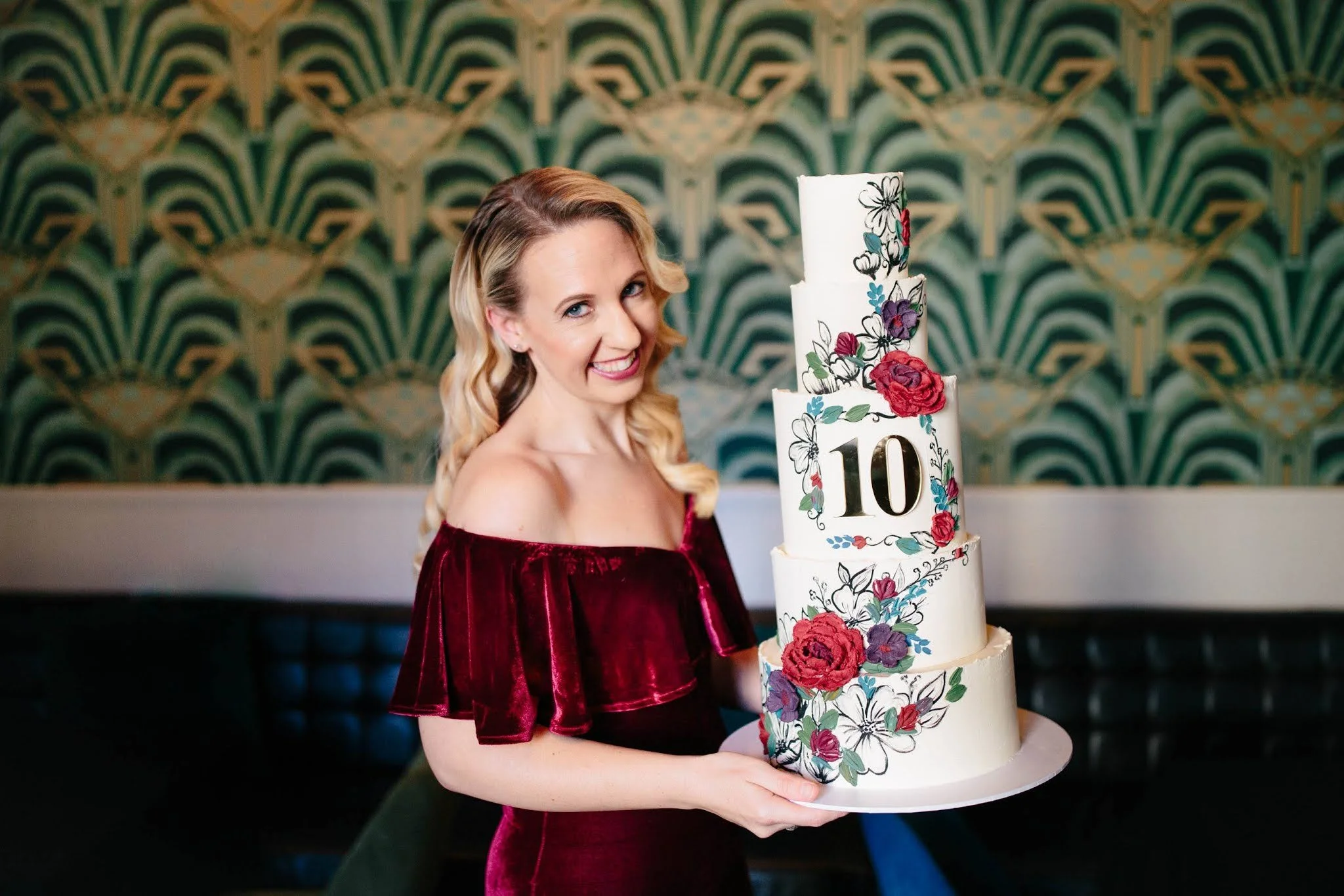 A woman with blonde hair in a burgundy off-shoulder dress holding a large, tiered birthday cake decorated with colorful flowers and the number 10, smiling at the camera.