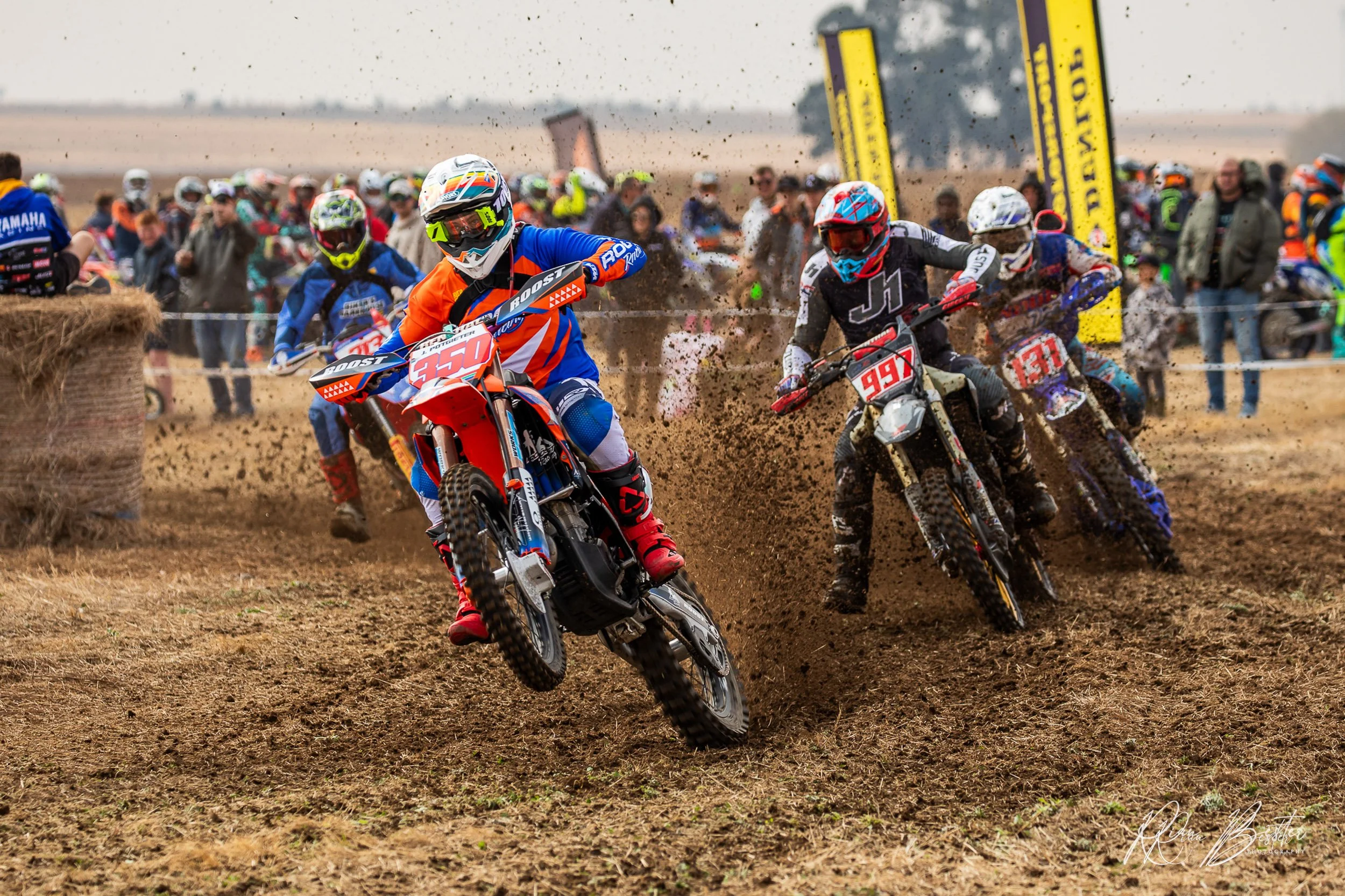 Motocross racers in colorful gear riding dirt bikes on a muddy track, kicking up dirt and in front of a crowd of spectators.