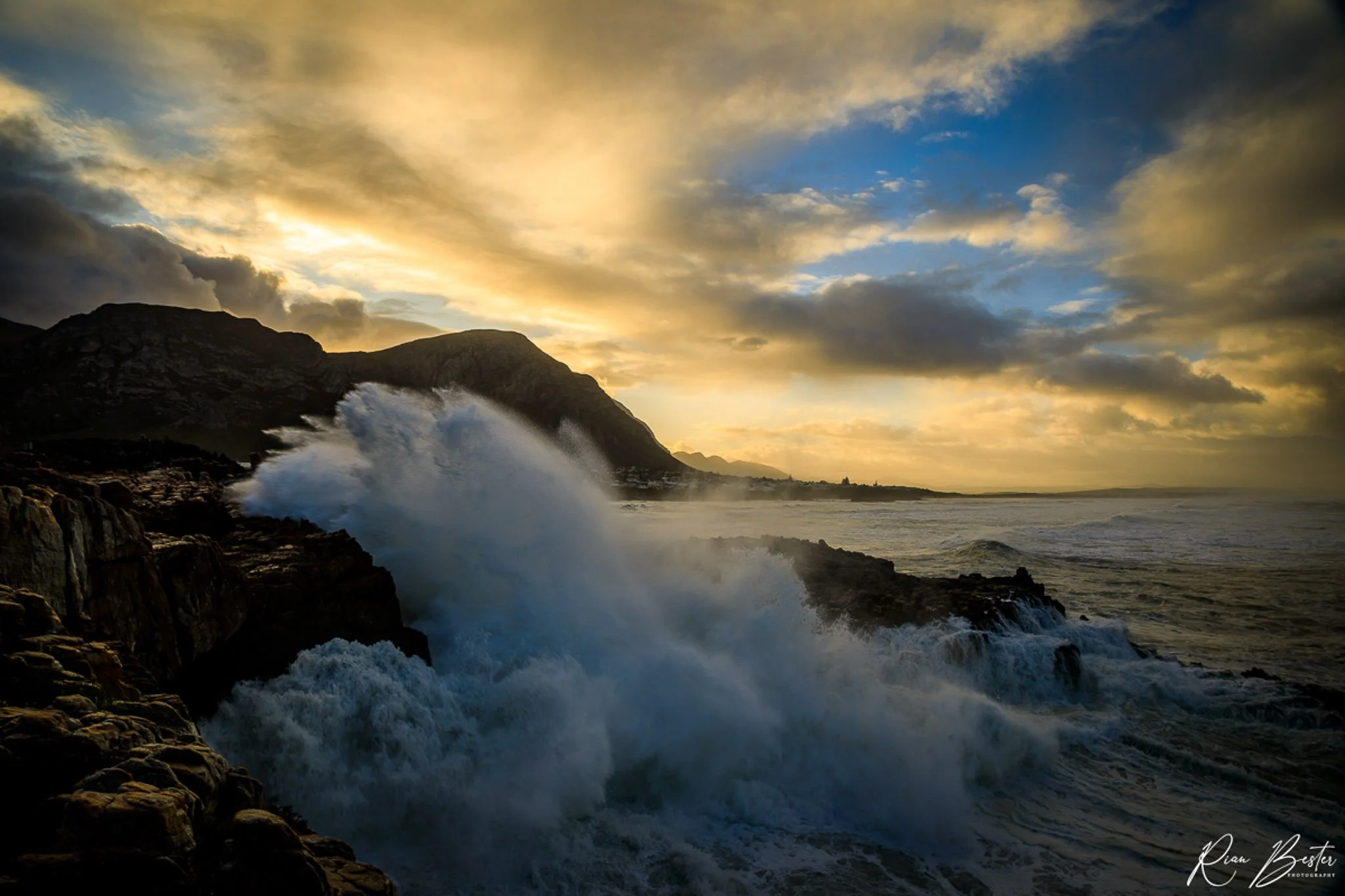 Ocean waves crashing against rocky shoreline during sunset with cloudy sky and mountain in background.