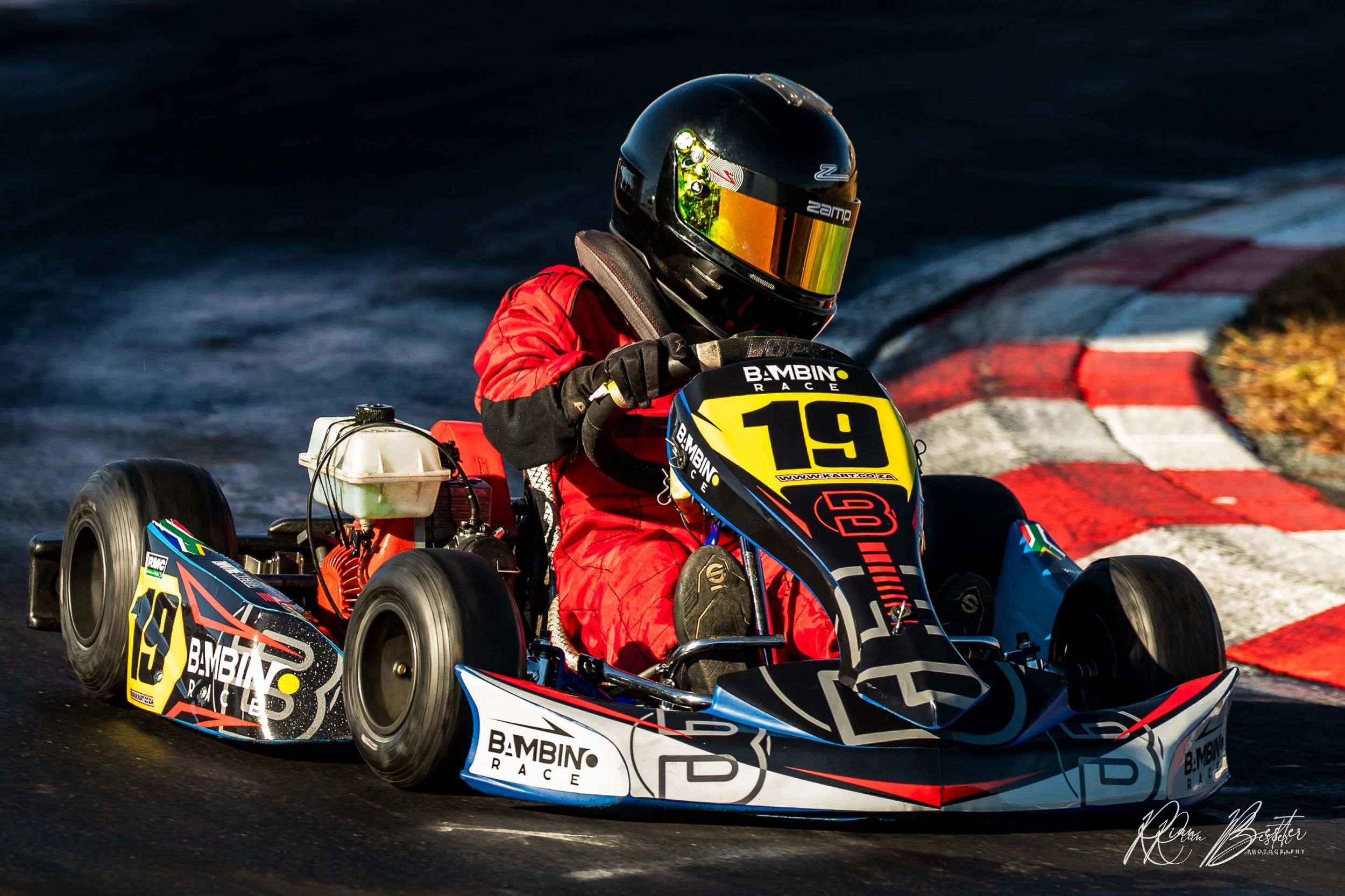 Go-kart driver in red racing suit and black helmet, navigating a turn on a race track, with red and white curbing visible.