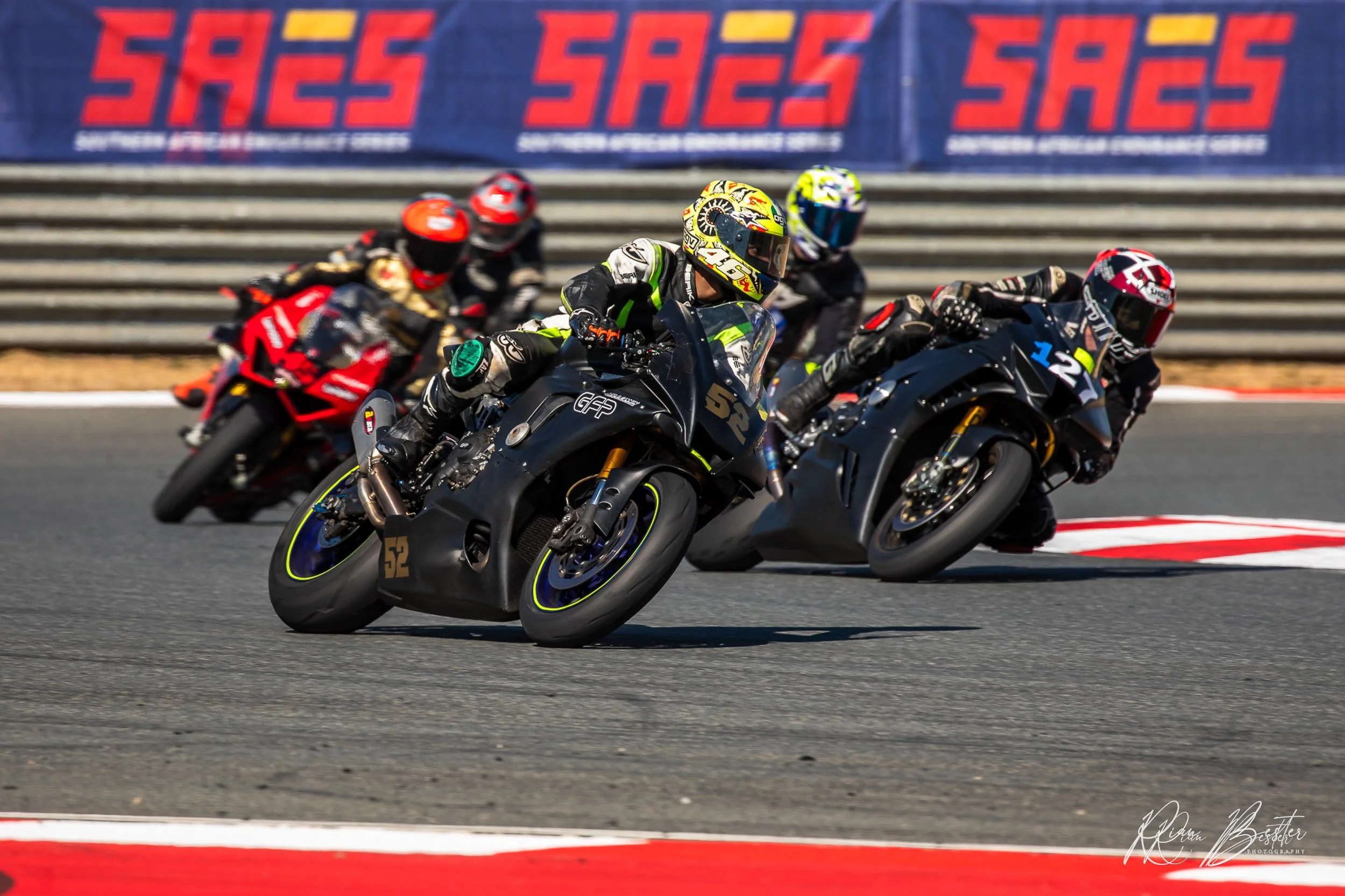Six motorcycle racers in racing gear leaning into a turn on a racetrack during a race, with a blue barrier in the background featuring the logo 'SAES'.