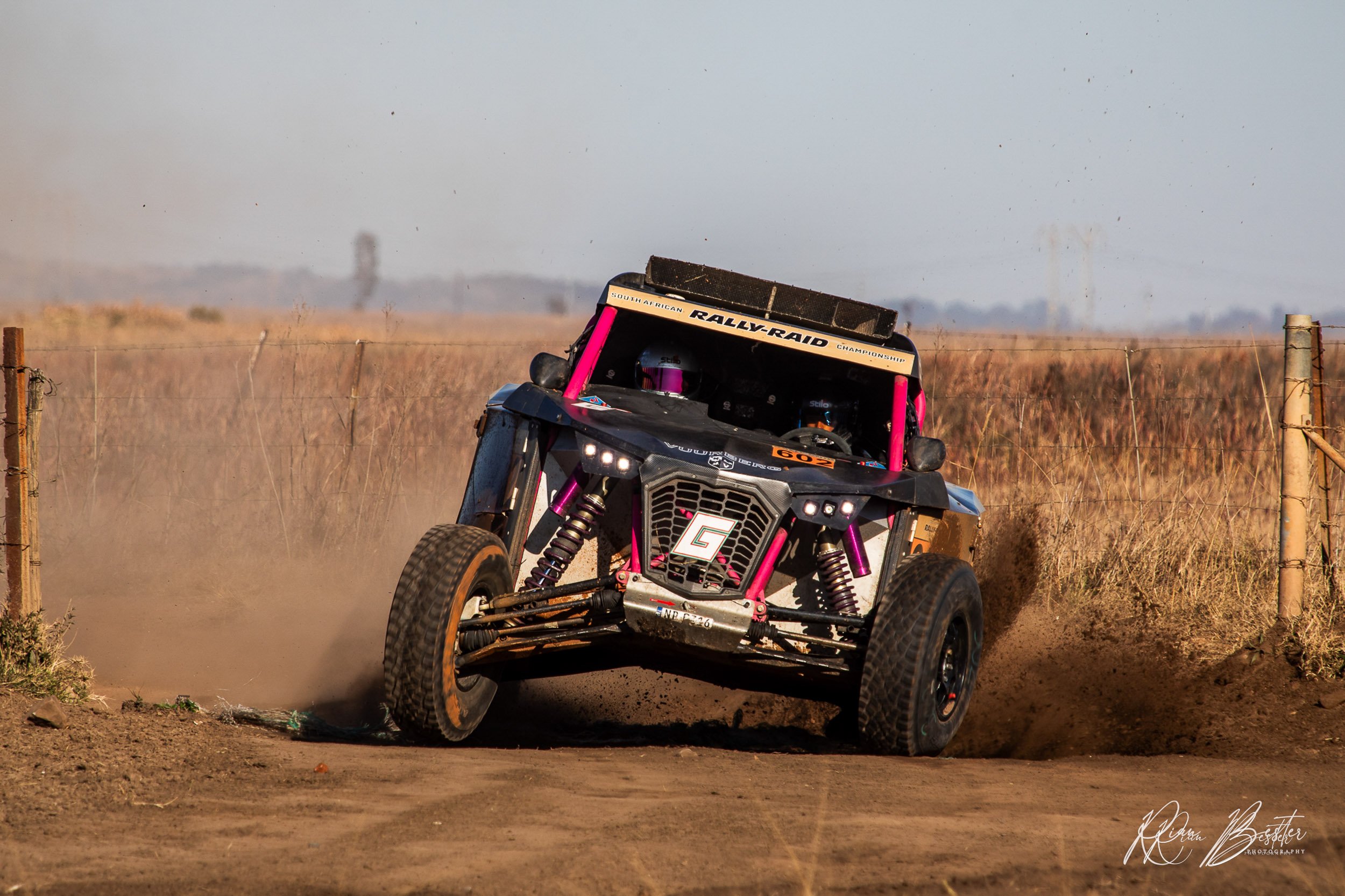 Off-road rally car kicking up dirt on a dusty track with a fence and open landscape in the background.