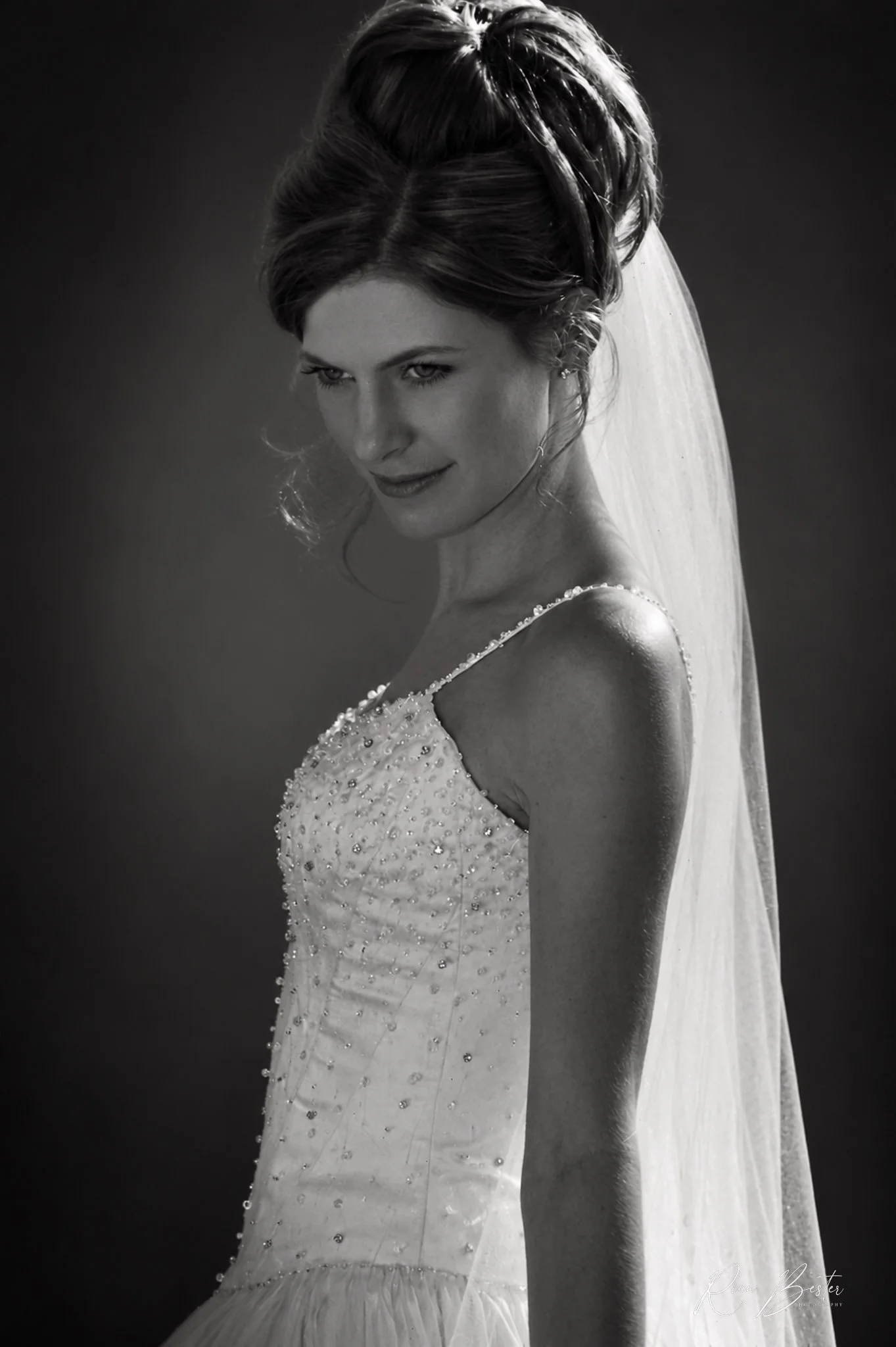 Black and white photo of a woman in a wedding dress, looking over her shoulder with a smile and a veil flowing behind her.