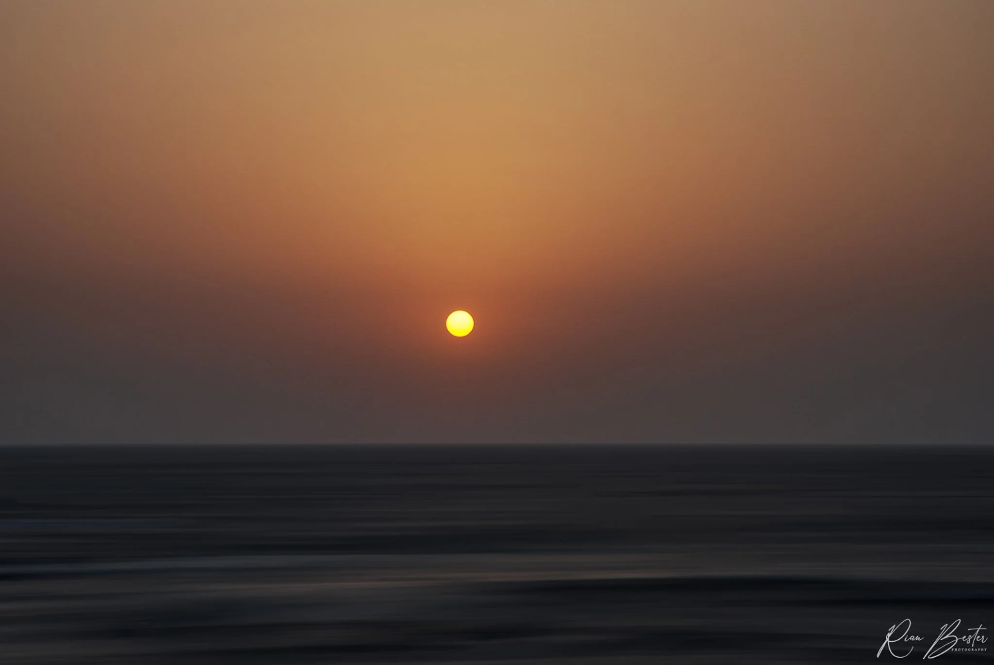Sun setting over the ocean with a colorful sky transitioning from orange to pink, and blurred waves in the foreground.