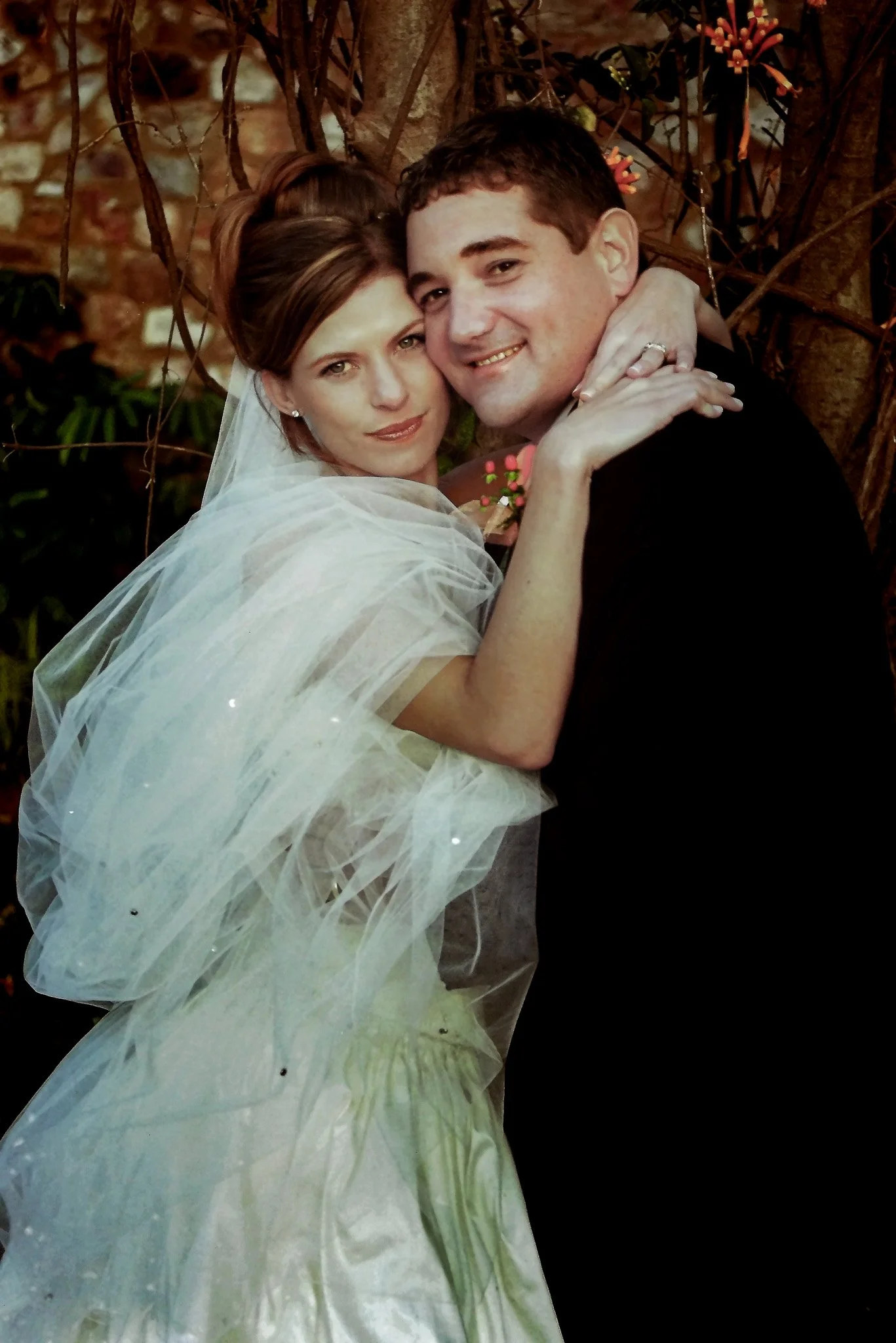 A newlywed couple smiling, with the bride wearing a sheer gown and veil, and the groom in a black suit, embracing outdoors near a tree with flowers and a brick wall in the background.